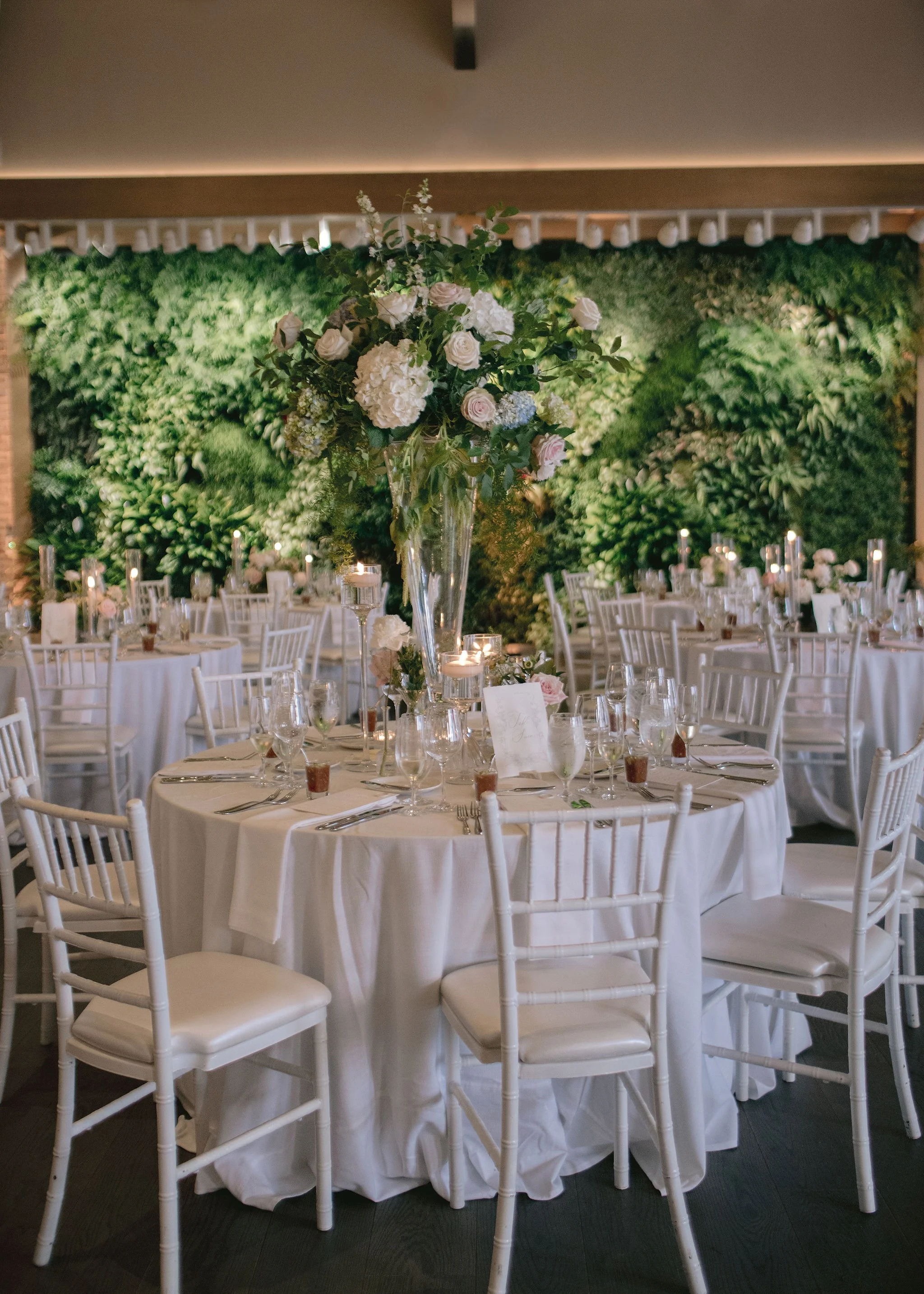 Elegant banquet hall decorated for wedding reception with round tables draped in white tablecloths, topped with floral centerpieces, candles, and glassware, set against a lush green wall.