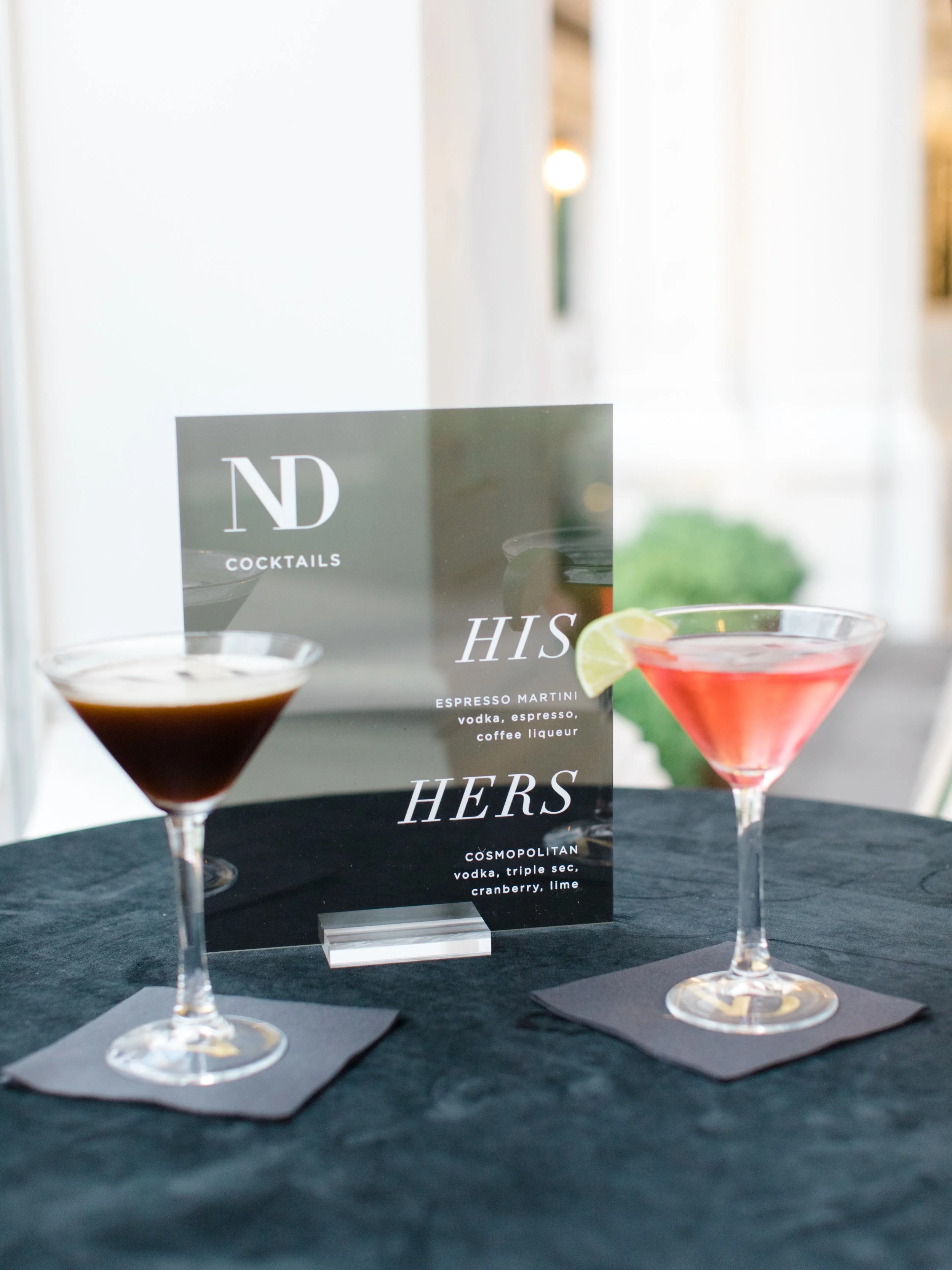 Two cocktails, one dark and one pink, on a black table with a menu card between them, featuring drinks with vodka, triple sec, cranberry, lime, espresso, vodka, espresso, and coffee liqueur.