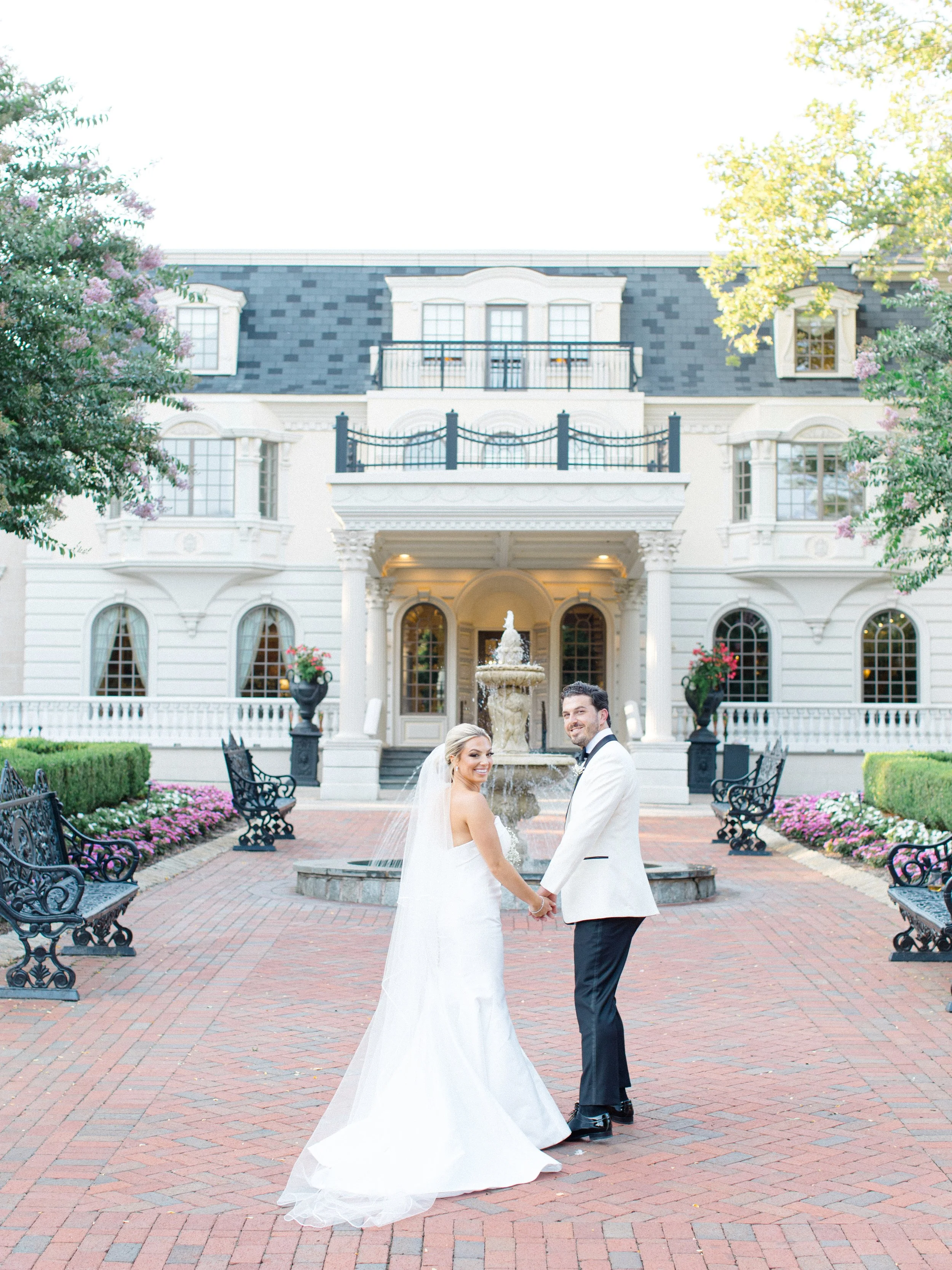 A bride and groom holding hands in front of a fountain outside a grand white wedding venue with trees and benches.