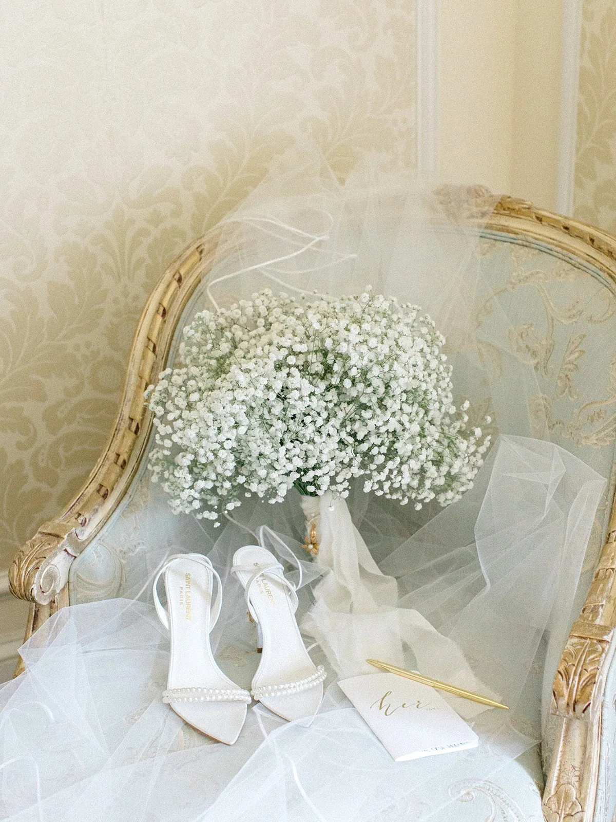 A pair of white high-heeled shoes with pearl embellishments on the ankle straps, a bouquet of white baby's breath flowers, a gold pen, and a small card with the word 'her' written in gold, all arranged on a vintage-style wooden chair with decorative 