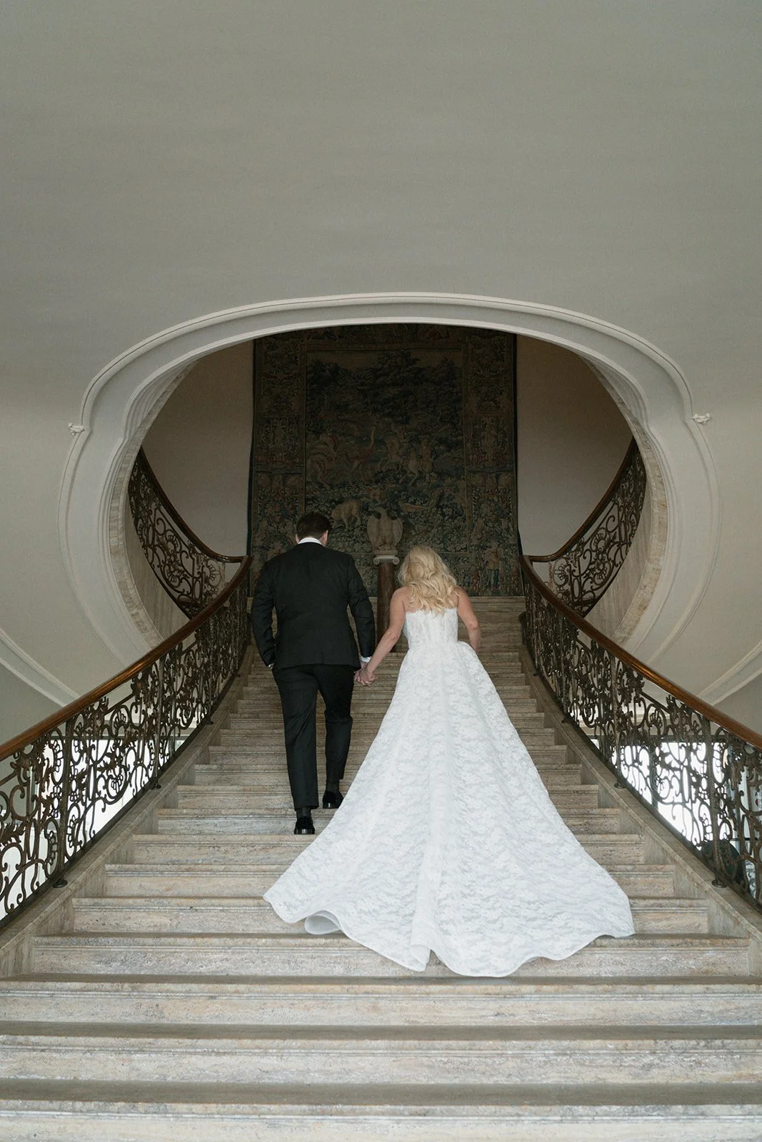 A bride and groom holding hands as they ascend a grand staircase, with ornate iron railings and a decorative tapestry at the top.