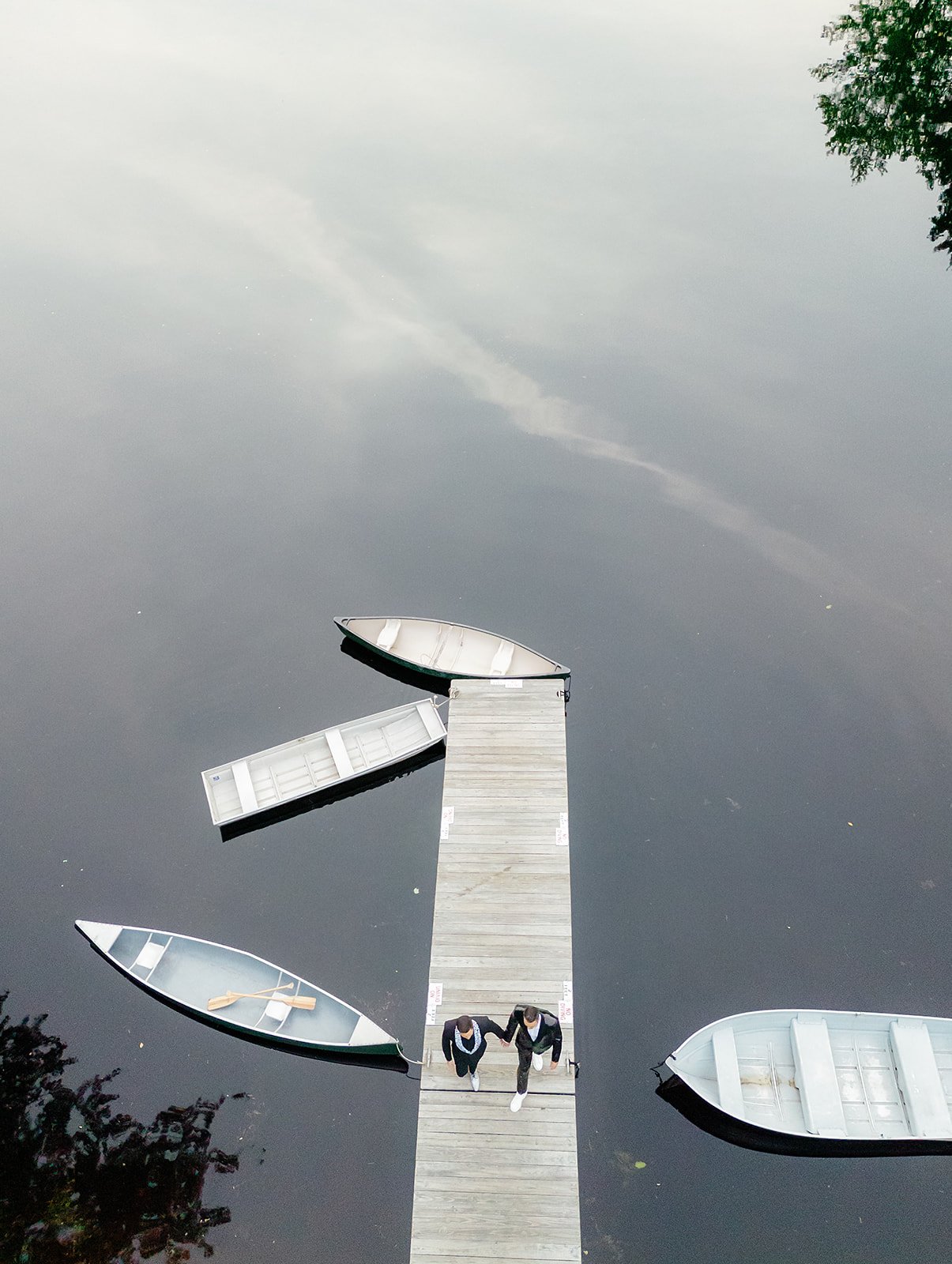 Two people in suits holding hands walk on a dock over a calm body of water with four boats, one tied to the dock and three floating nearby. The water reflects the cloudy sky and trees.