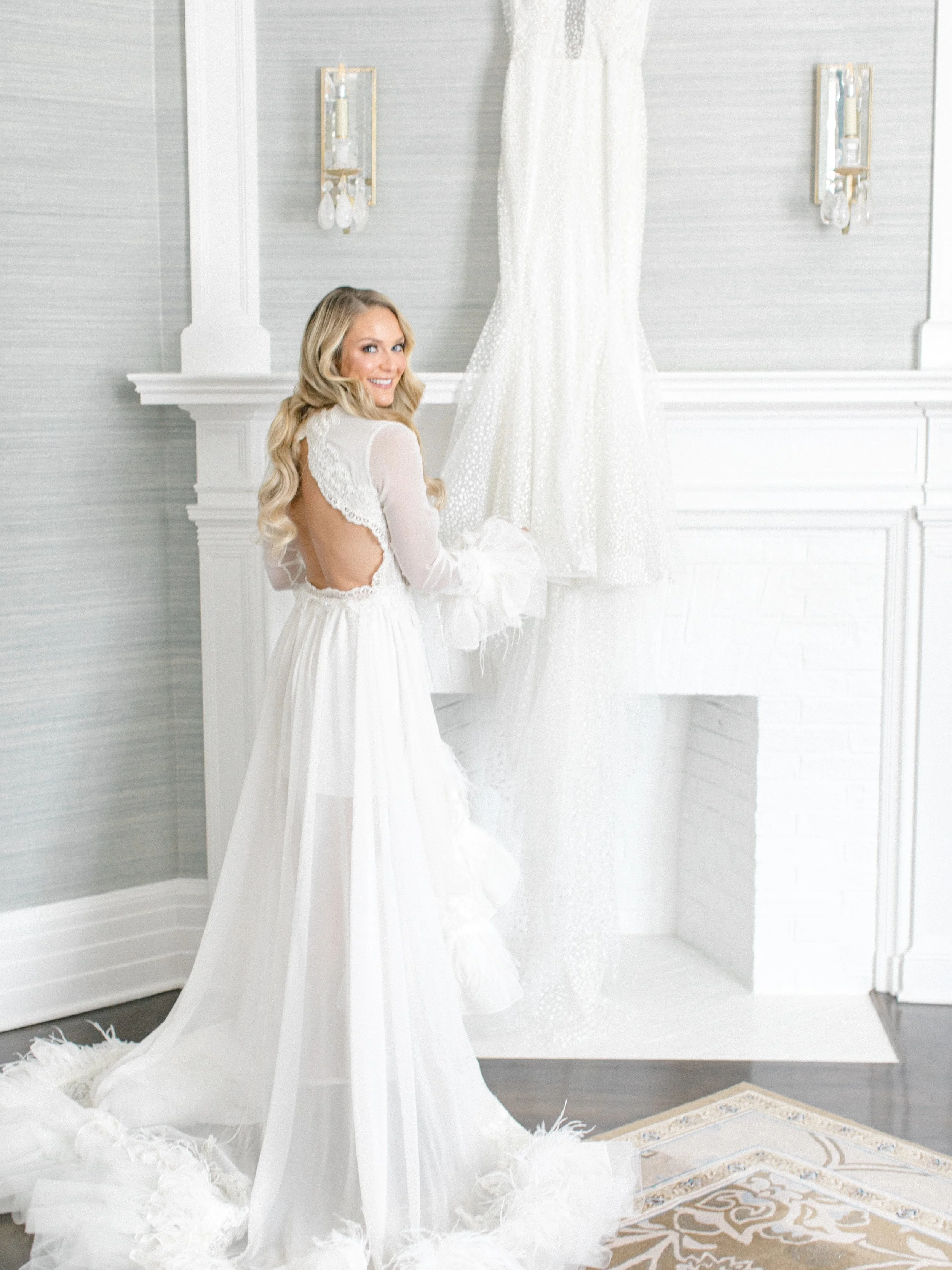 A woman in a white wedding dress smiling and holding a lace veil in front of a white fireplace with a mannequin dressed in another bridal gown