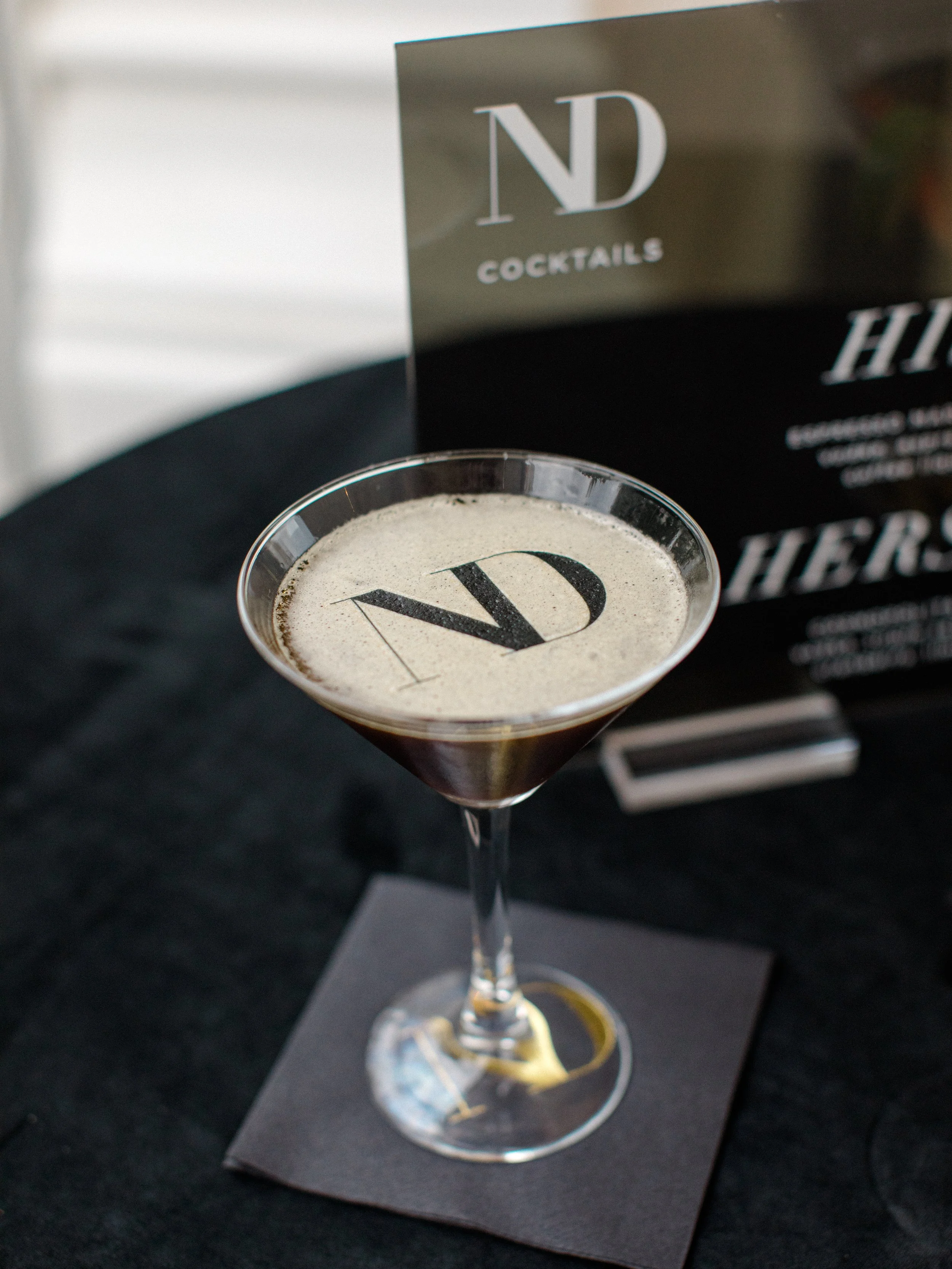 A cocktail glass with a dark drink topped with foam, featuring the letter 'V' on the foam, placed on a black napkin on a black table, with a black sign promoting ND Cocktails in the background.