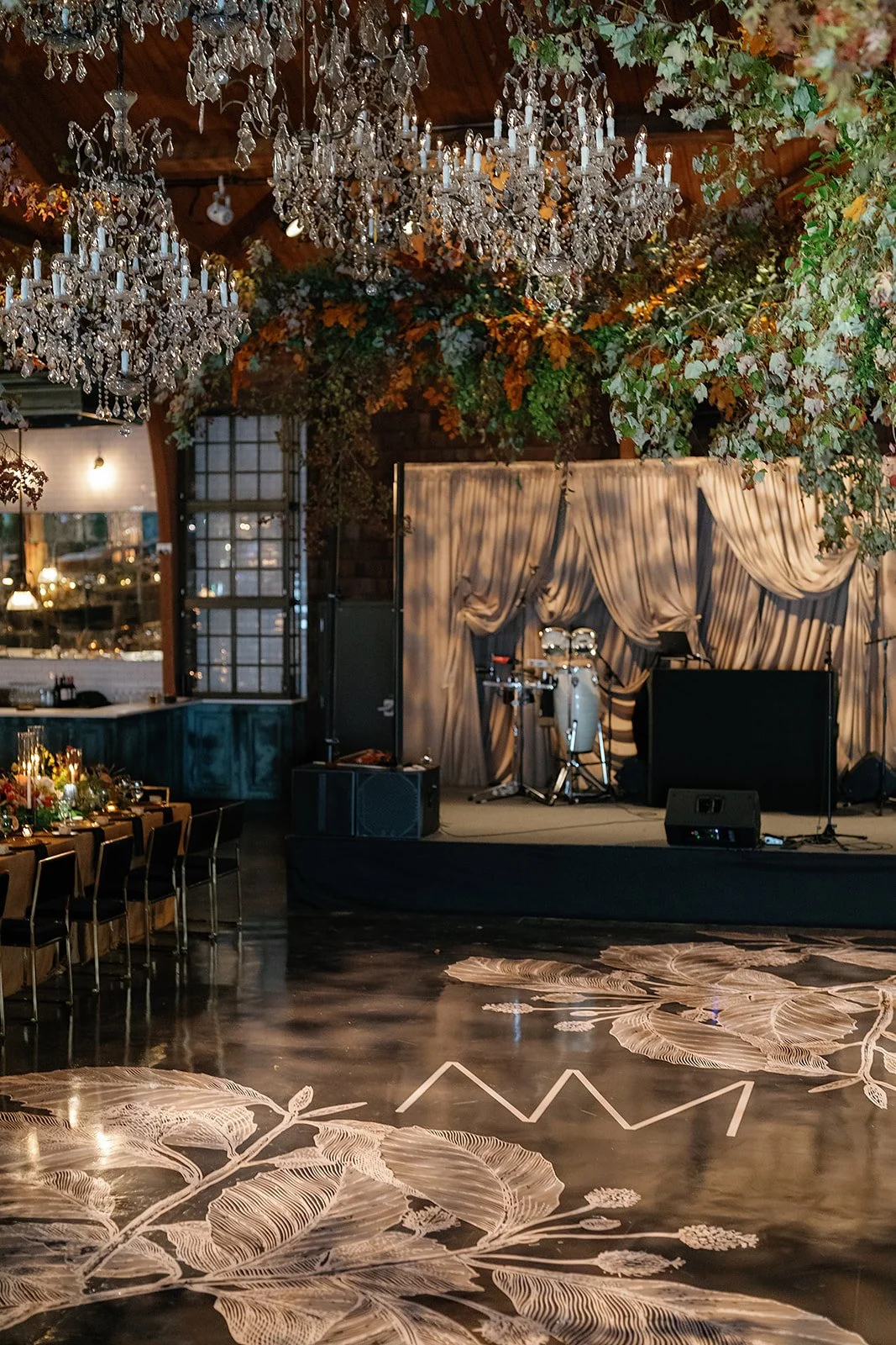 Elegant event venue with a stage decorated with draped curtains, musical instruments, and a sound system. The ceiling features hanging crystal chandeliers and lush greenery with flowers, and the floor has artistic floral patterns and initials 'MM'.