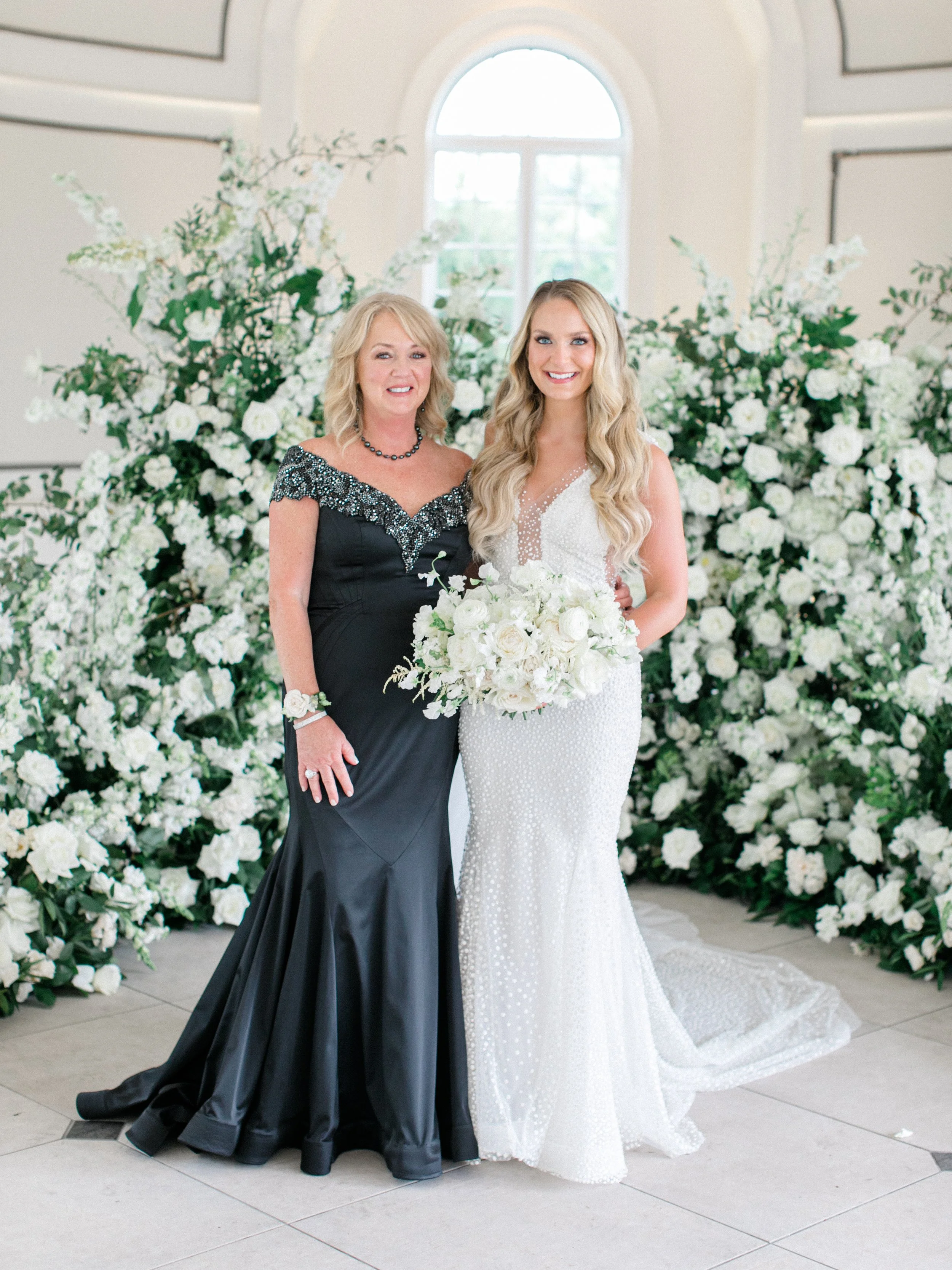 A bride and another woman stand together in front of lush white flowers and greenery, indoors near a large arched window, both smiling. The bride wears a white beaded wedding gown and holds a white bouquet, while the other woman wears a black dress w