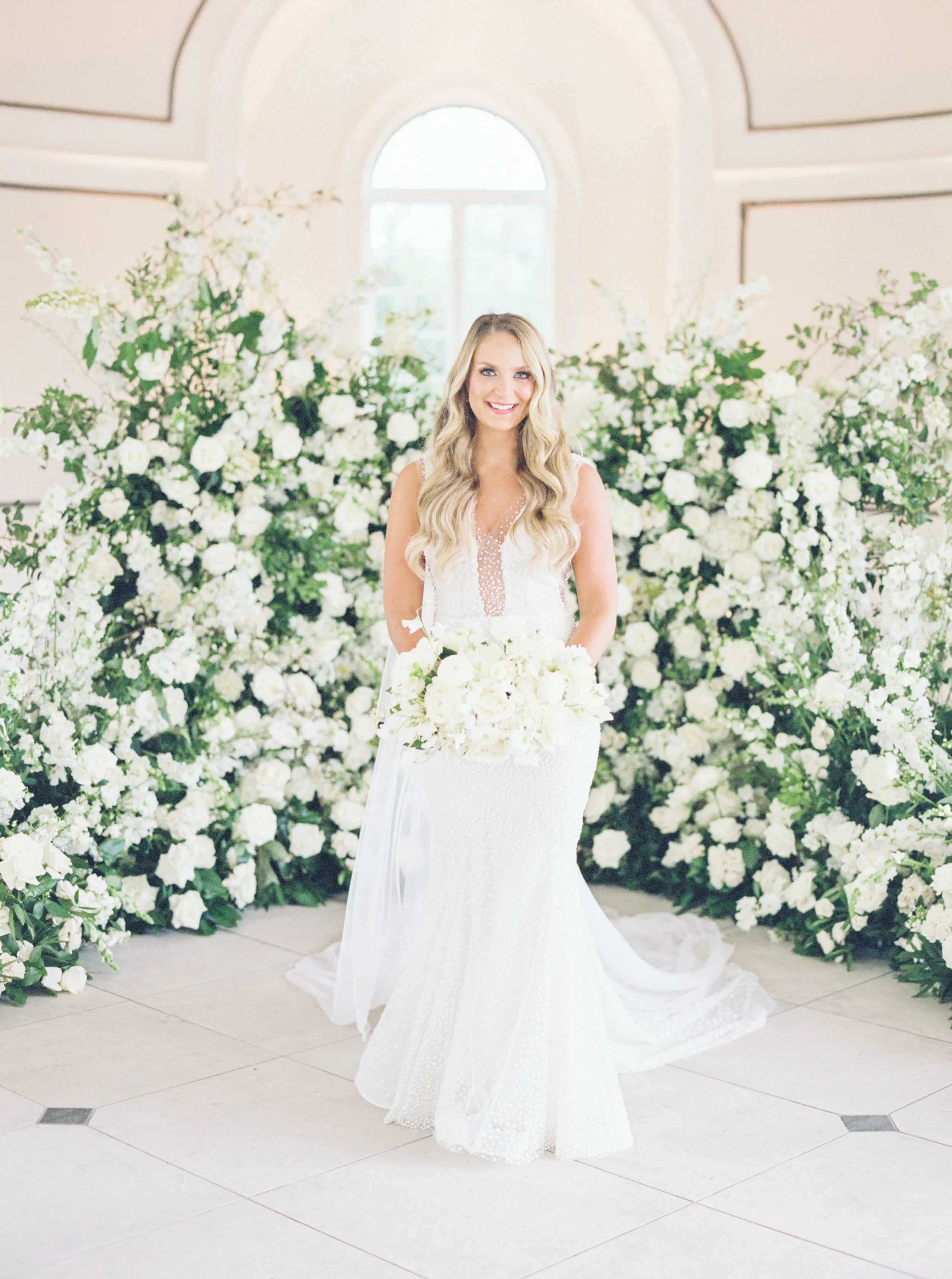 A bride in a white wedding dress holding a bouquet of white flowers, standing in front of an elaborate backdrop of white flowers and greenery, with a large arched window in the background.