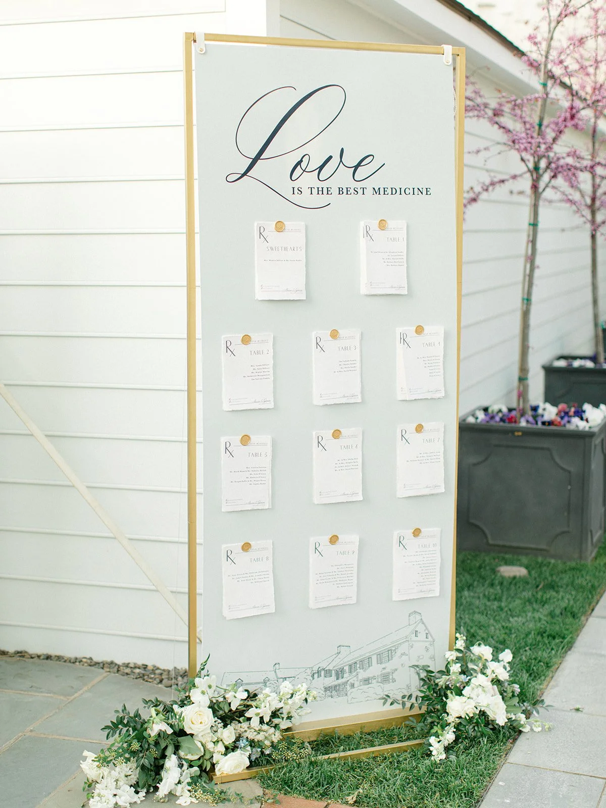 A white wedding seating chart board with the quote "Love is the best medicine" at the top, decorated with white flowers at the base, outside near a house with white siding and flowering trees.