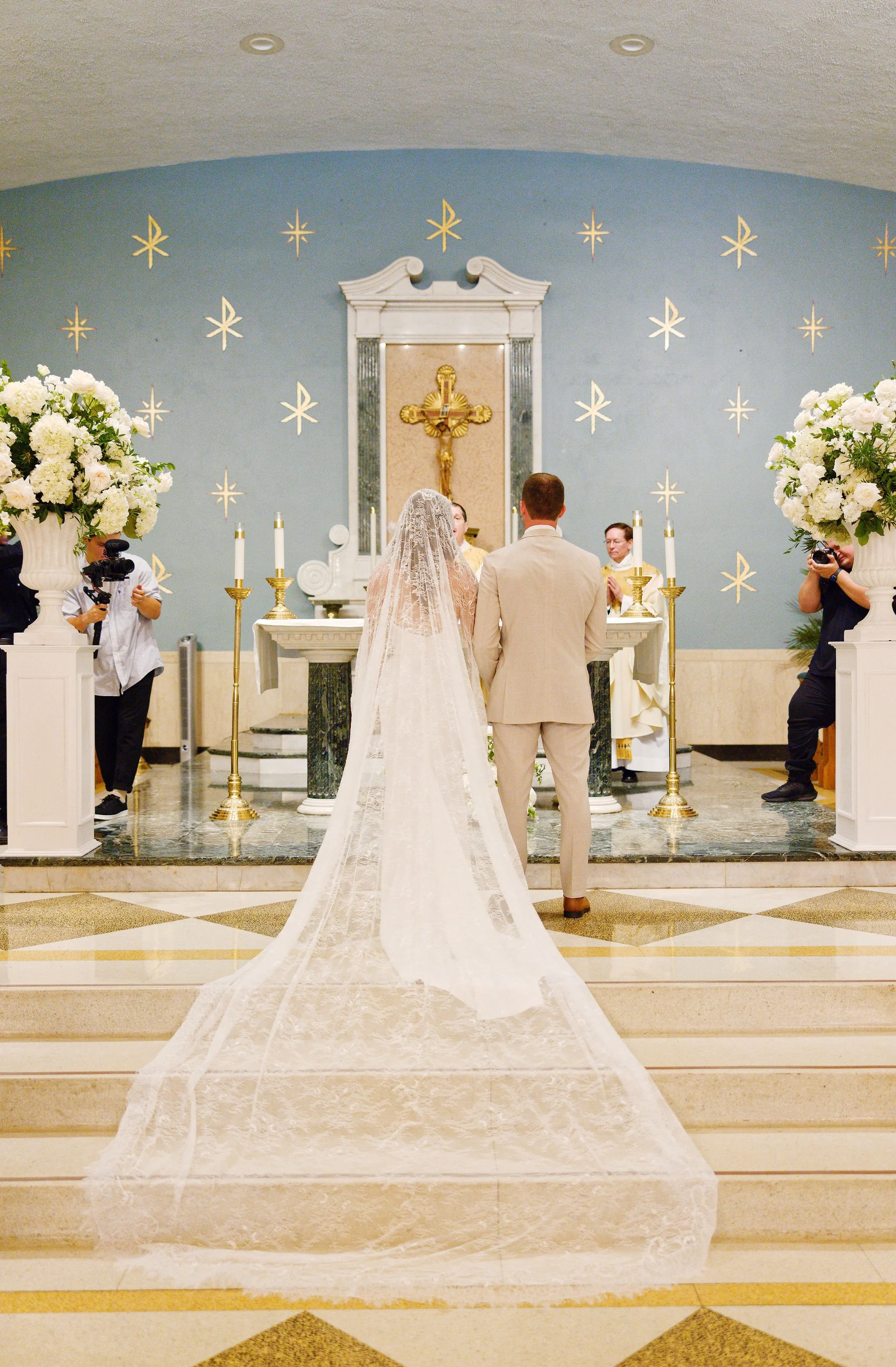 A wedding ceremony taking place inside a church, with the bride and groom standing at the altar facing the officiant, surrounded by floral arrangements and baptismal candles.