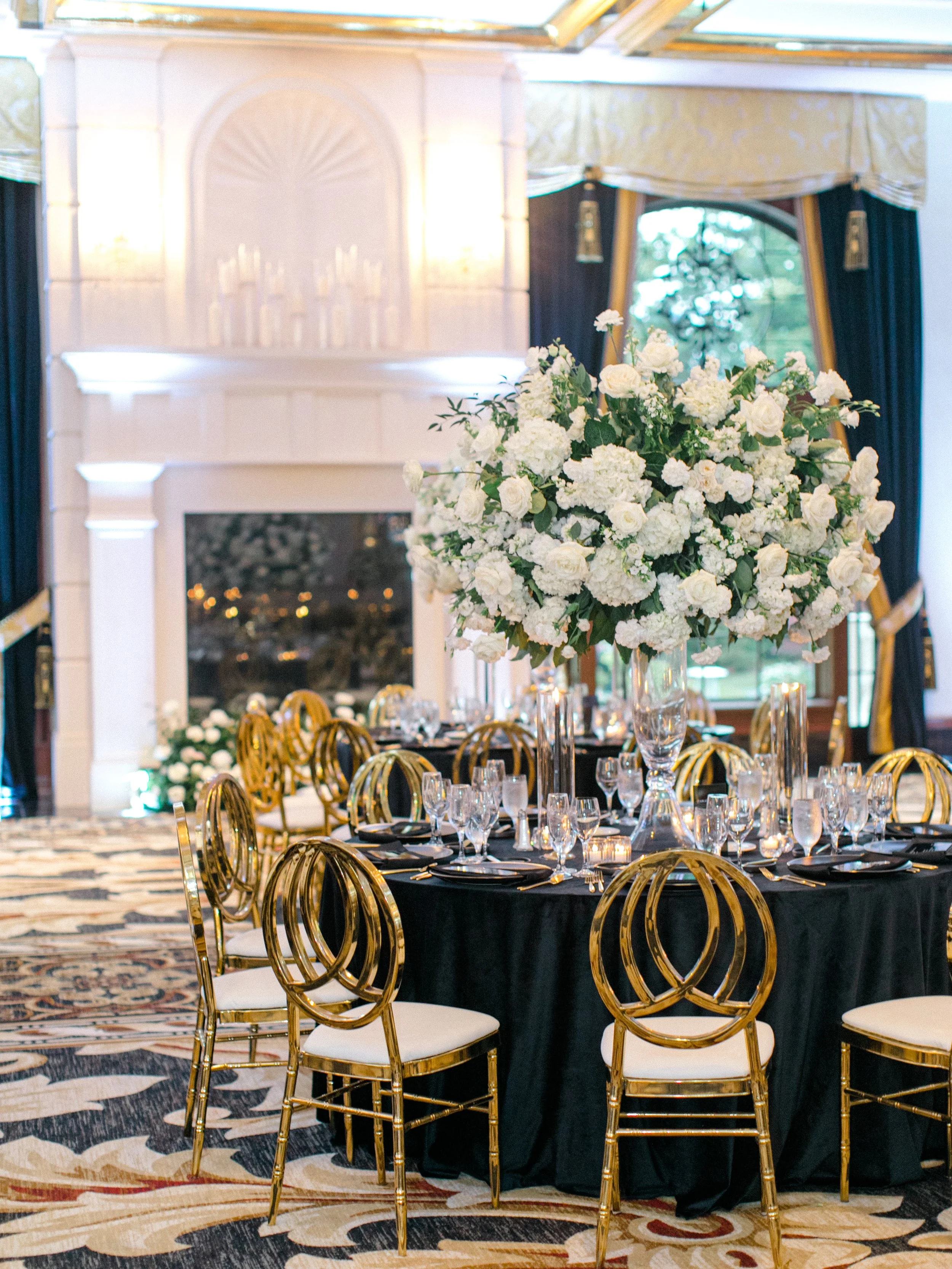 Elegant banquet table with a large floral centerpiece of white roses and hydrangeas, gold chairs, black tablecloth, and assorted glassware in a decorated event space with a fireplace and large windows.