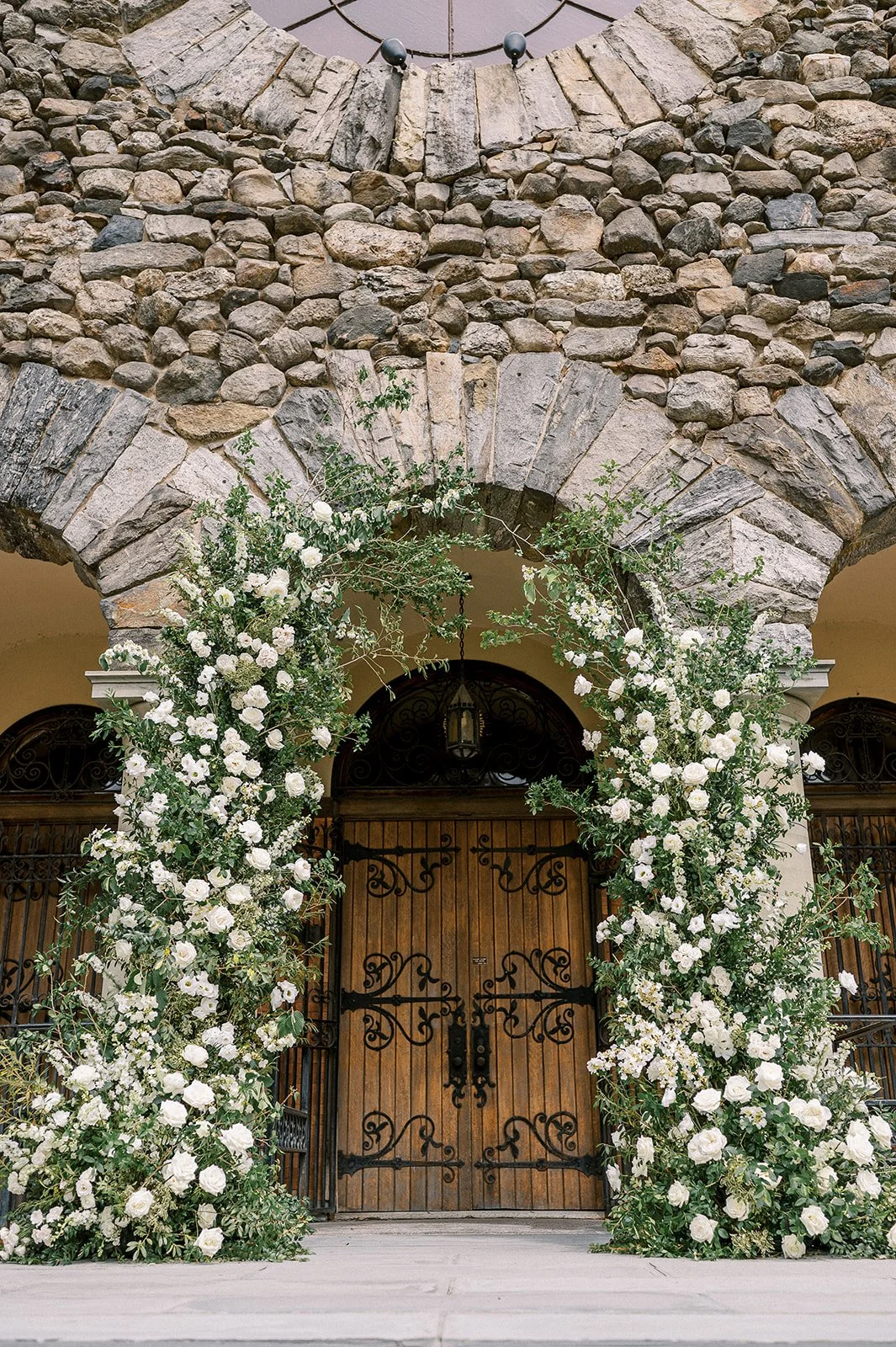 Wedding arch with white flowers and greenery in front of wooden double doors with black wrought iron detailing, stone wall above, hanging lantern above door.