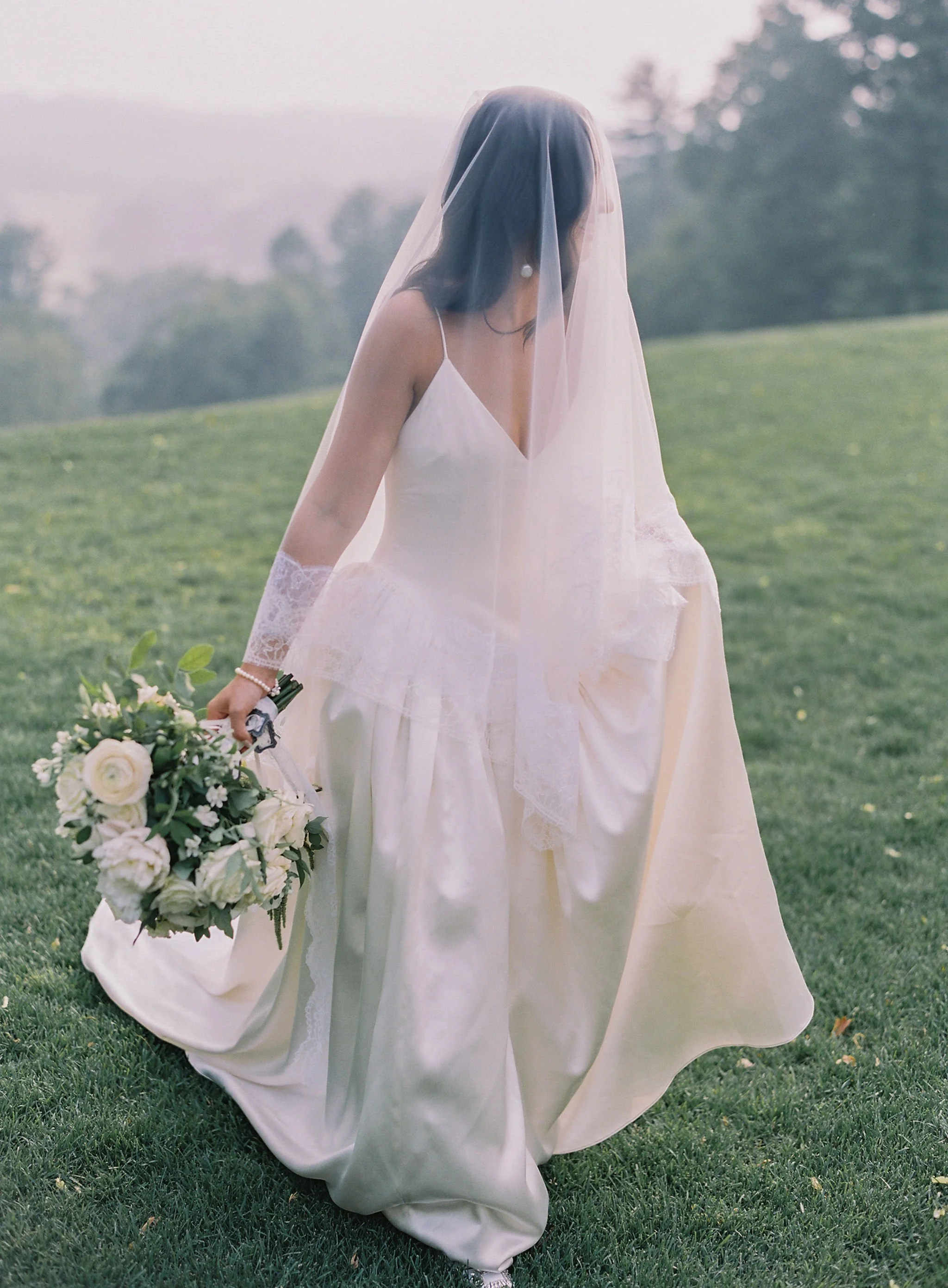A bride in a white wedding dress and veil holding a bouquet of white flowers, walking on a grassy field.