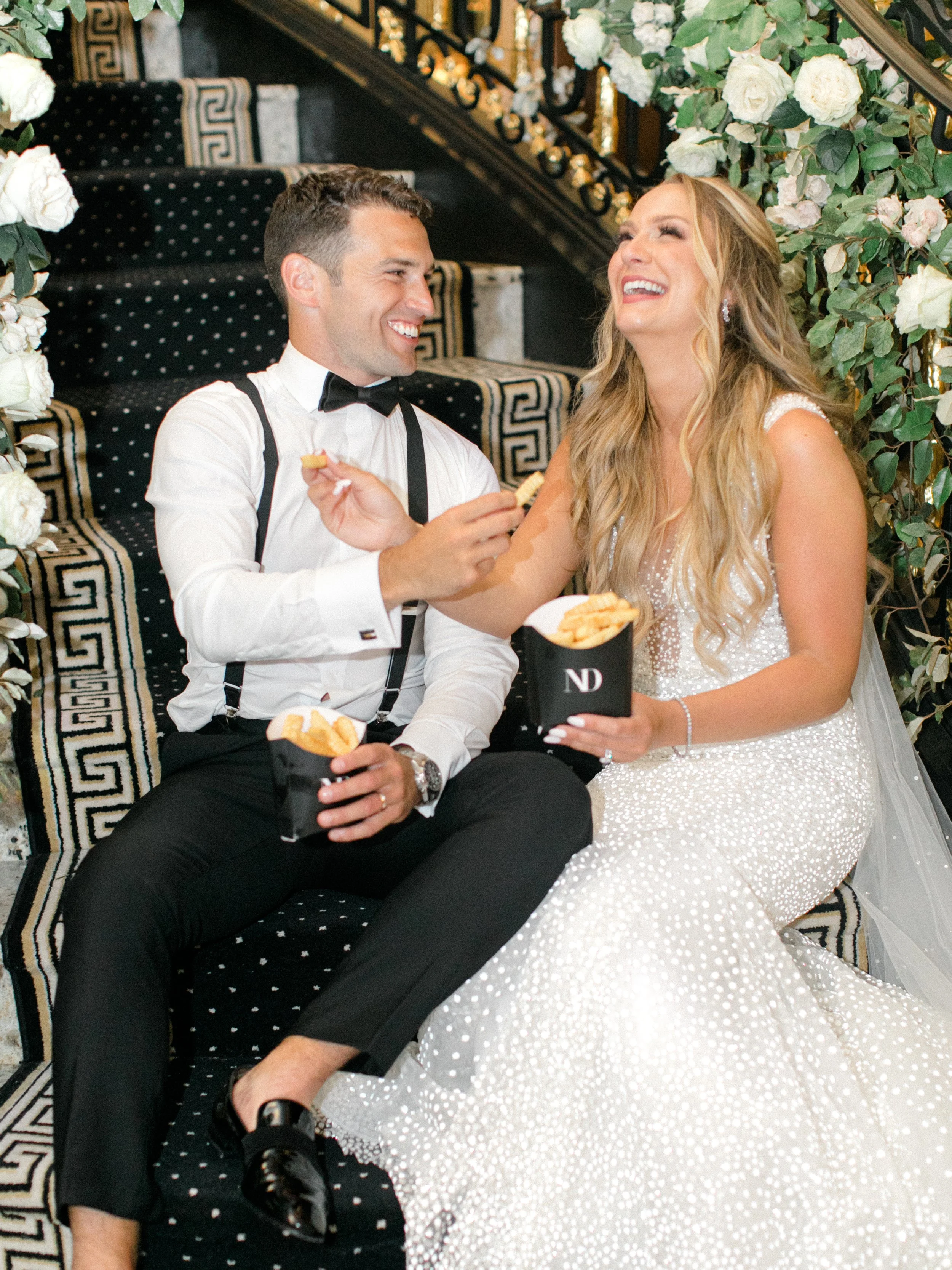 A bride and groom sitting on a black and gold staircase, enjoying French fries and smiling at each other, surrounded by white roses and greenery.