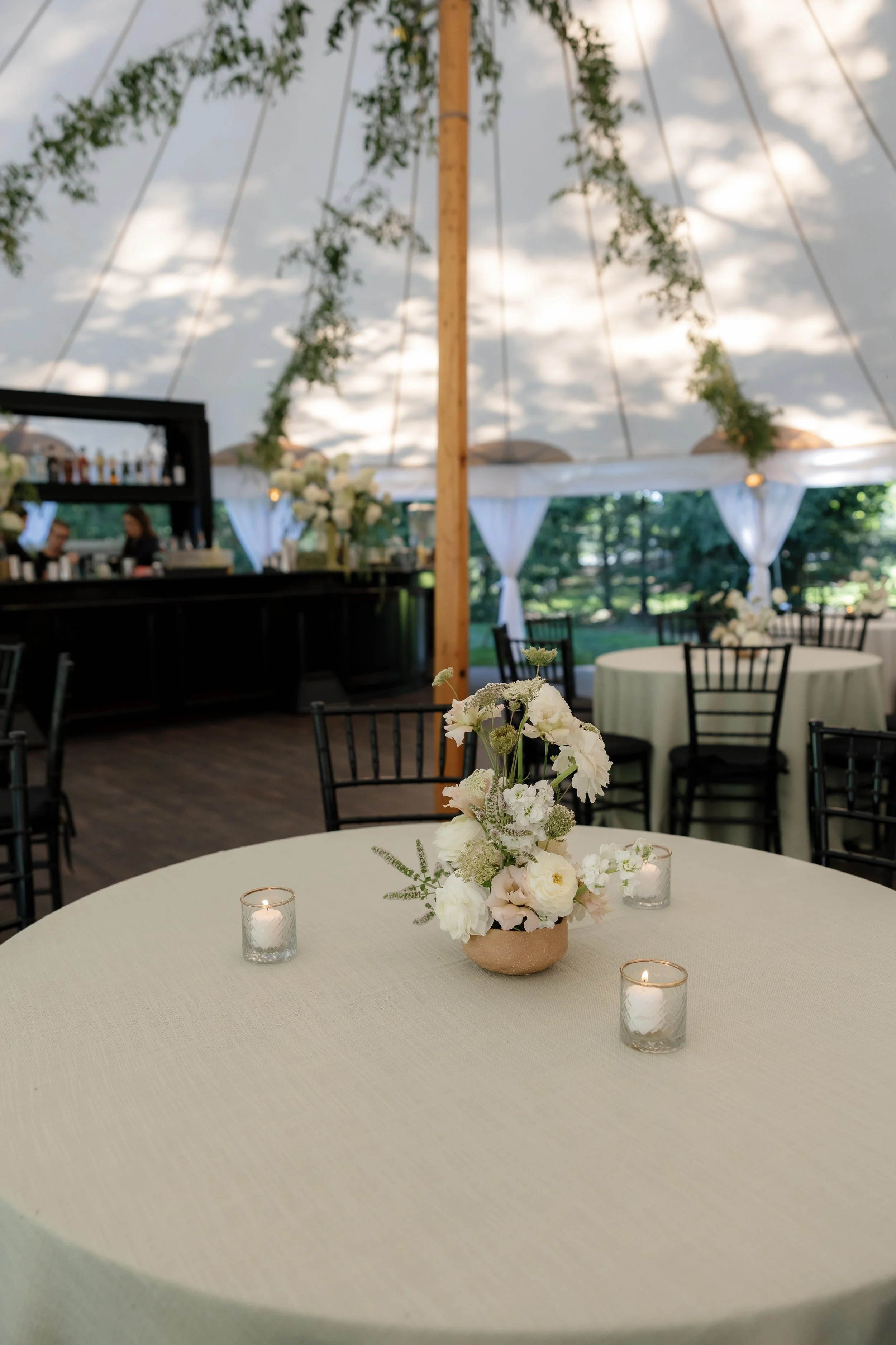 Elegant outdoor event setup with round tables, white tablecloths, floral centerpieces, and candles. A canopy with drapes and greenery decorates the space.