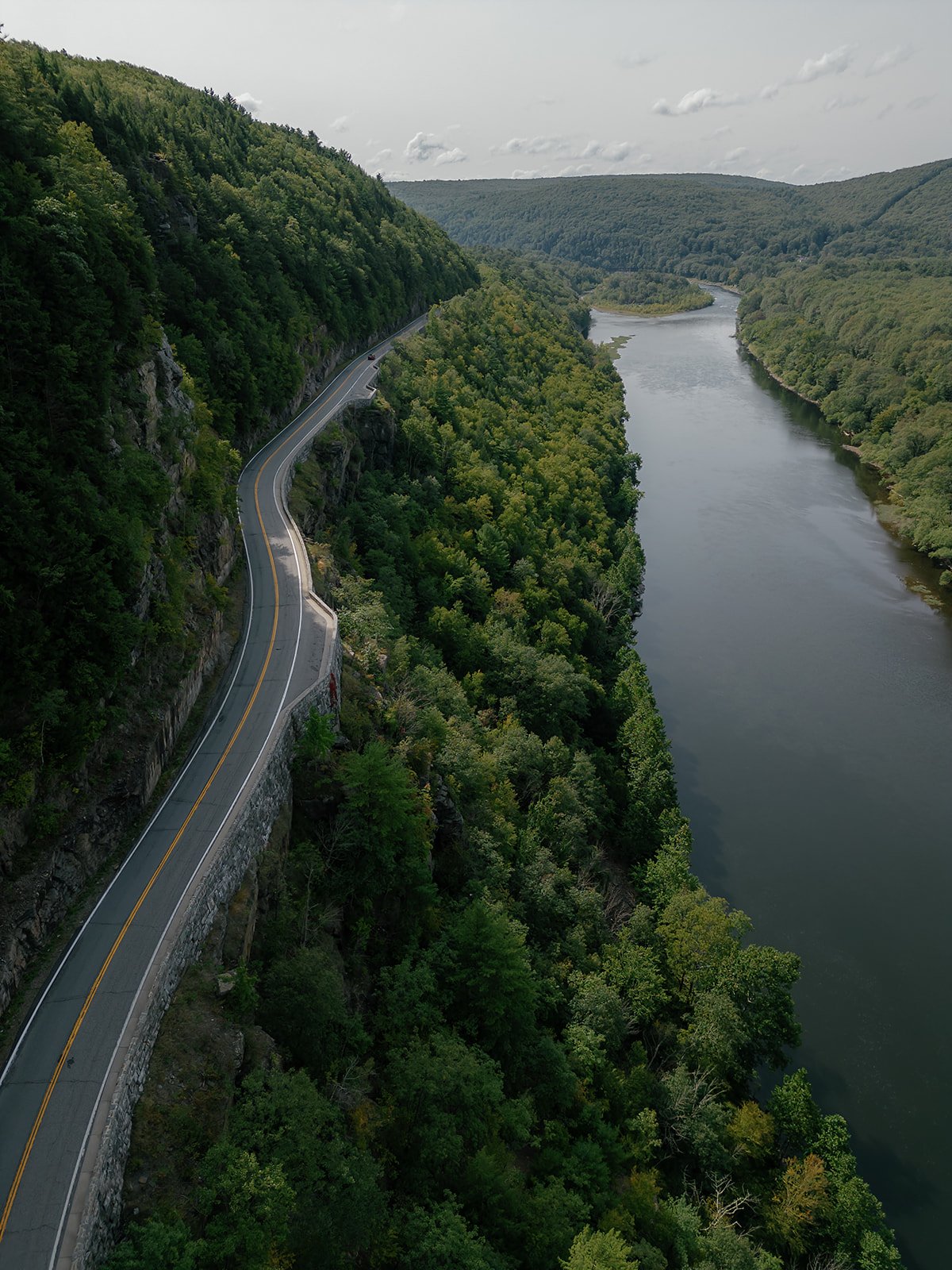 A winding road runs along a steep cliffside covered in dense green trees, with a river flowing parallel at the base of the cliff, surrounded by rolling hills covered in trees.