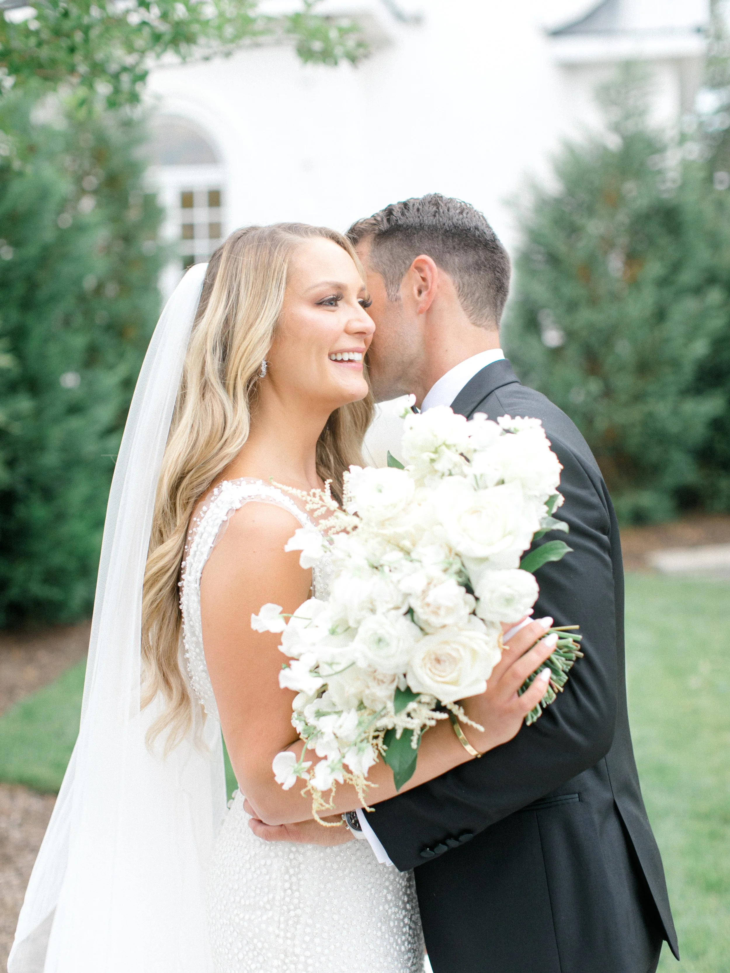A bride and groom sharing a moment outdoors, the bride holding a large white bouquet, smiling, wearing a white wedding gown, and the groom in a black tuxedo.