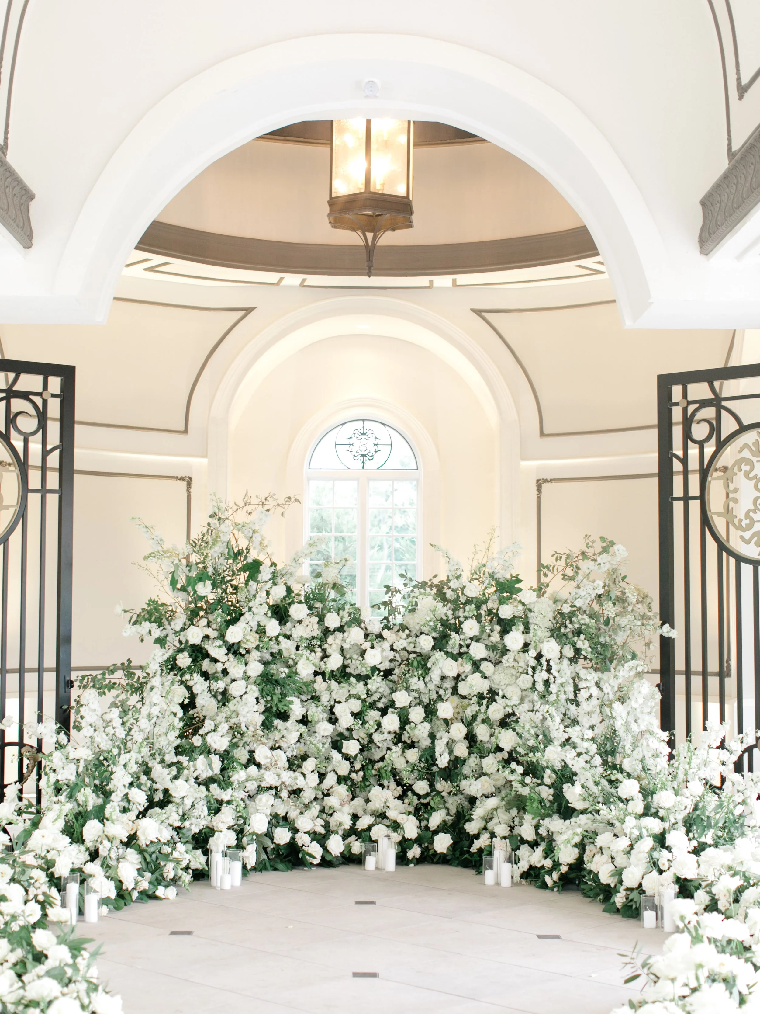 A wedding altar decorated with large arrangements of white flowers, candles, and greenery inside a bright, elegant room with arched windows and a lantern overhead.