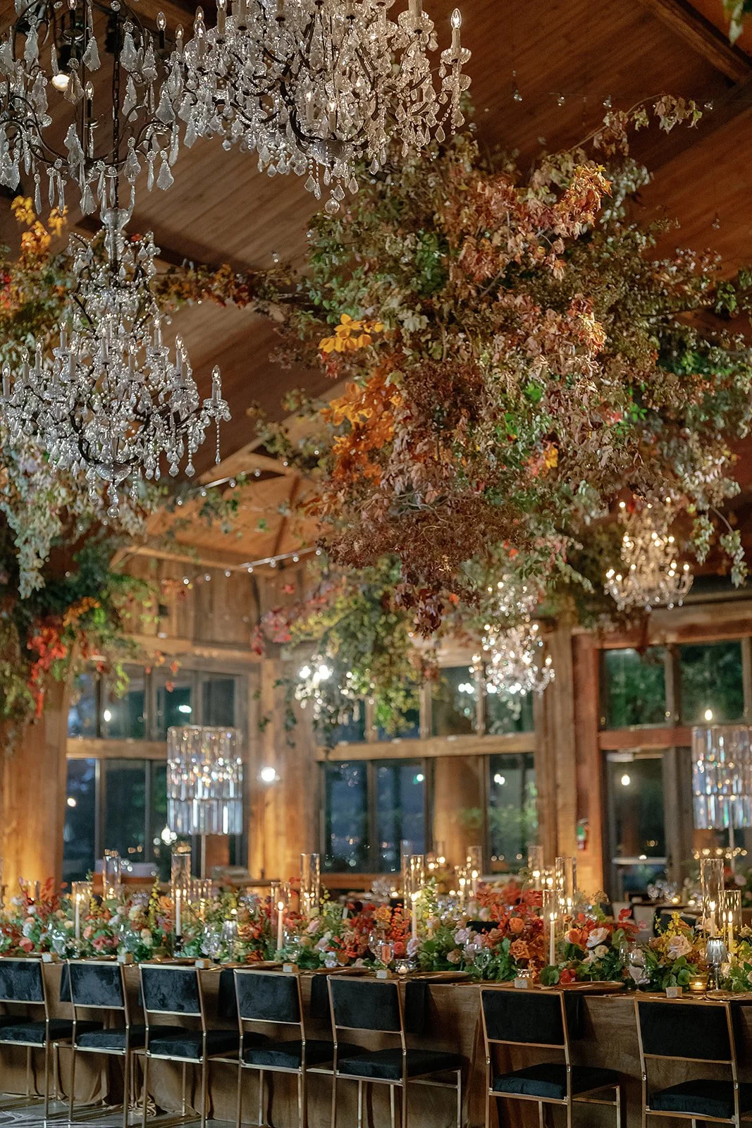An elegant event space featuring a long dining table decorated with colorful floral arrangements and candles, hanging greenery and flowers from the ceiling, large chandeliers, and wooden walls with large windows at night.