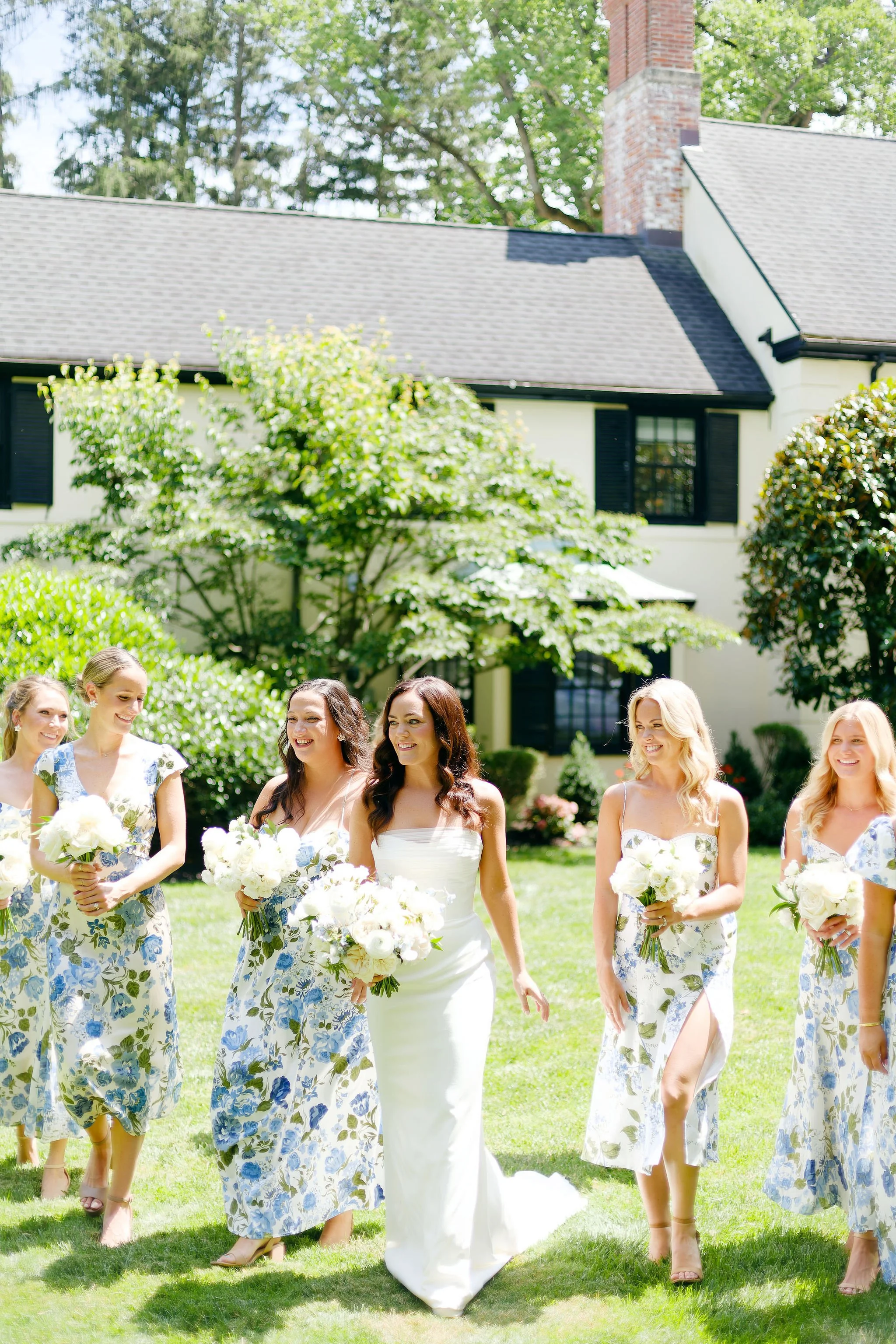 A bride and bridesmaids walking on a lawn in front of a house with trees in the background. The women are wearing white and blue floral dresses and holding bouquets.