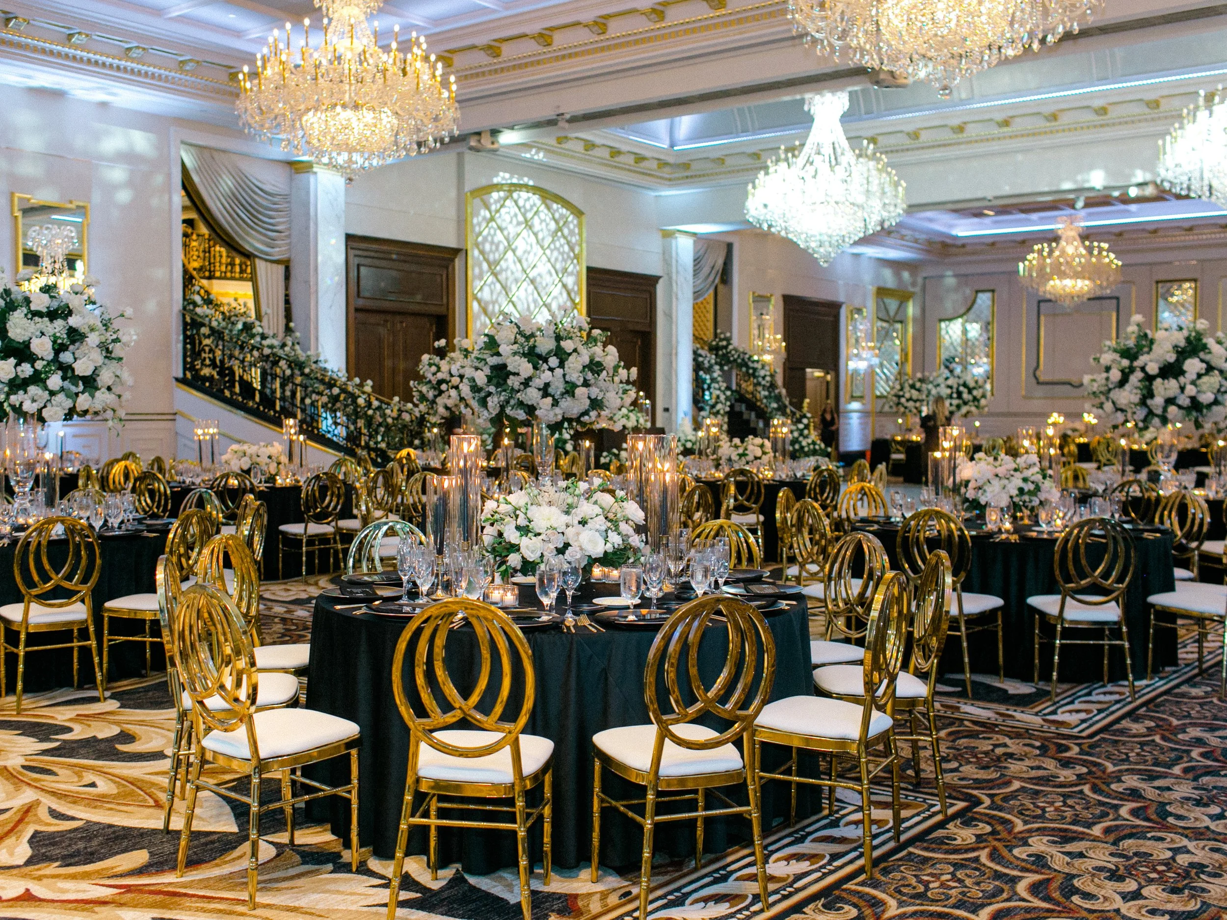 Elegant banquet hall decorated with white floral centerpieces and candles on black-draped round tables with gold chairs, chandeliers hanging from the ceiling, ornate moldings, and mirrors on the walls.