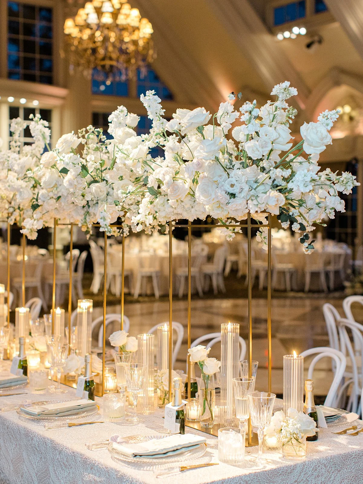 Elegant wedding reception table with tall gold floral centerpieces, white flowers, candles, and champagne bottles, in a decorated ballroom.