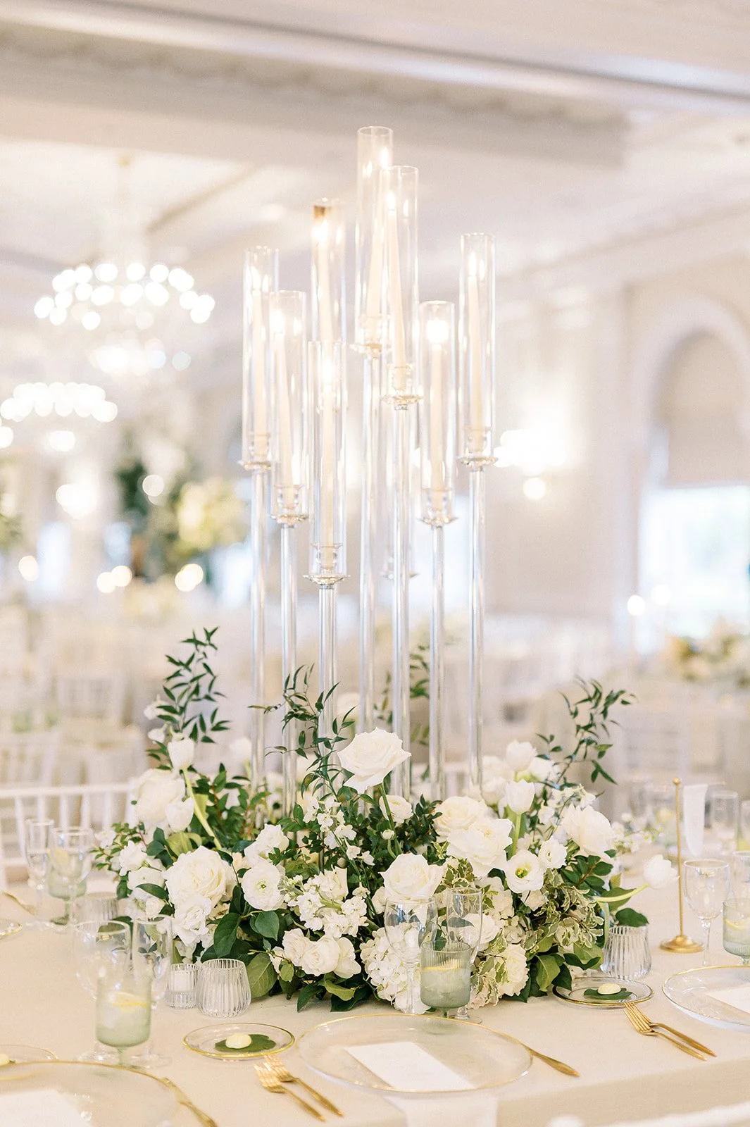 Elegant wedding table centerpiece with tall glass candle holders and white floral arrangement with green foliage.