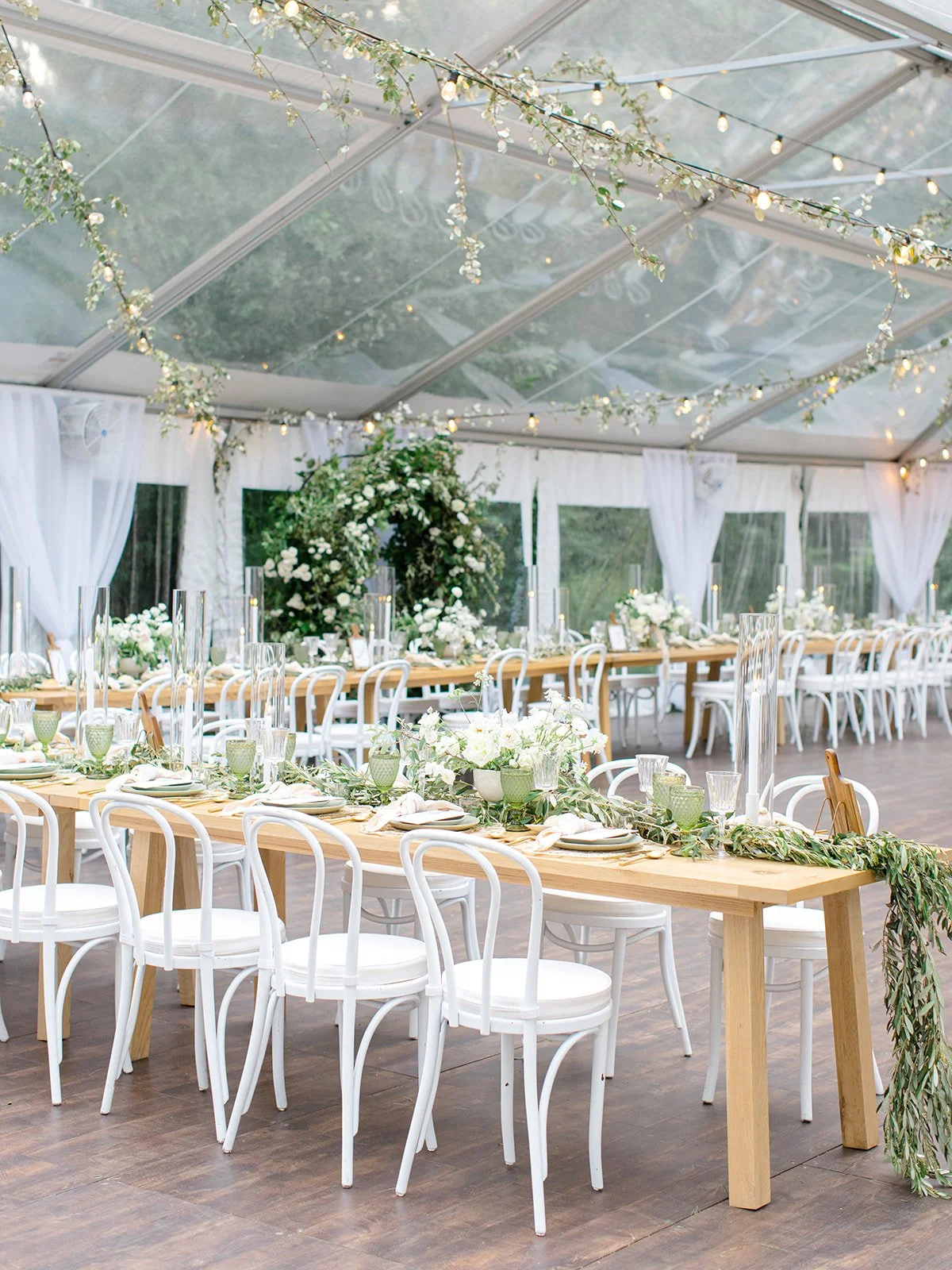 A decorated event space with long wooden tables and white chairs, adorned with white floral centerpieces and greenery. The space is covered with a transparent tent with string lights and hanging floral decorations.