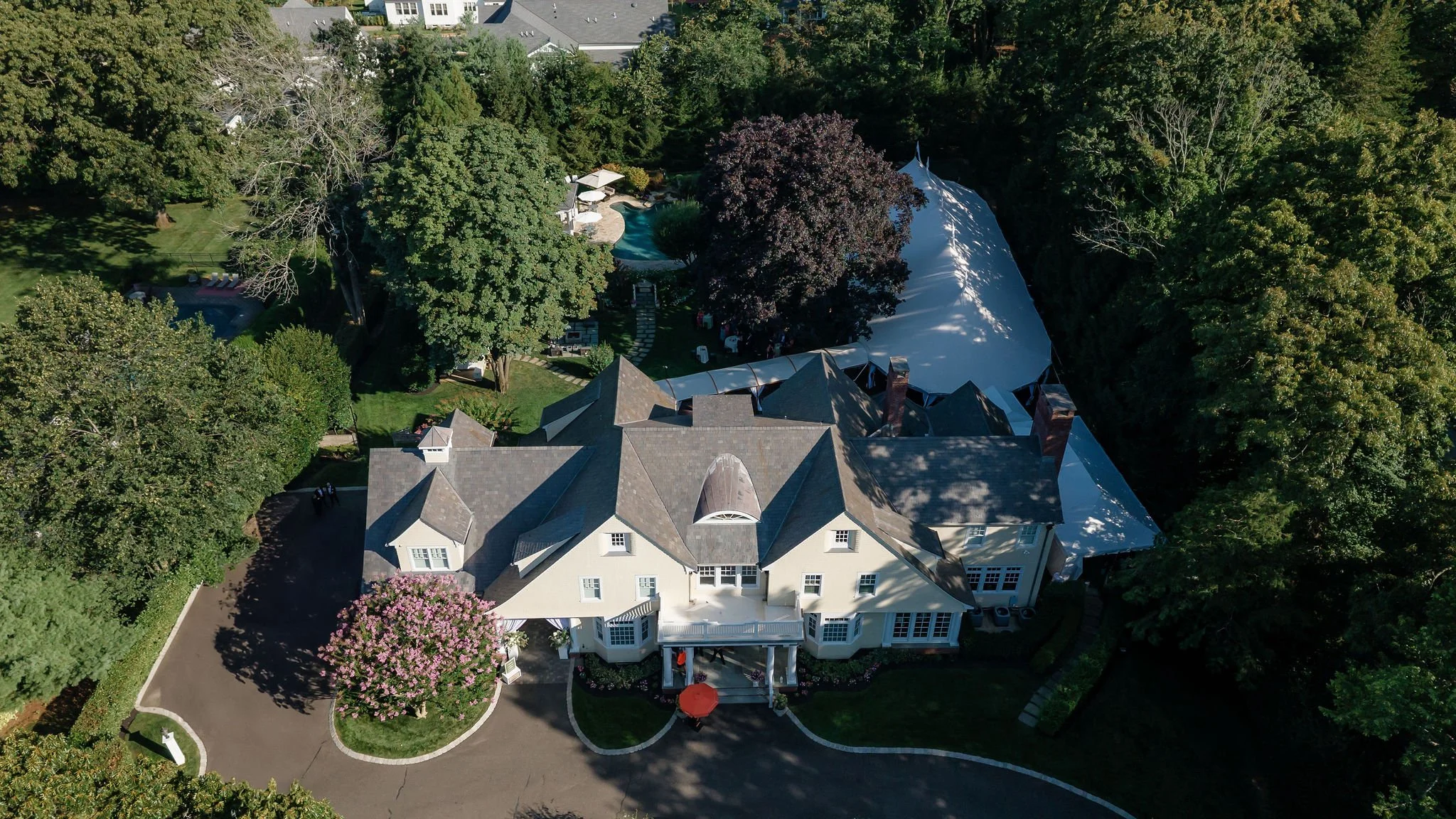 A large white house with a well-maintained lawn and a driveway, surrounded by tall green trees, with a large tent or event setup nearby and a swimming pool in the backyard.