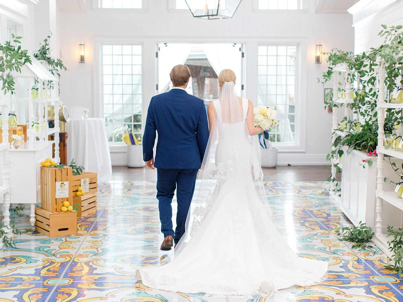 A bride and groom walking into a decorated indoor wedding venue, with the bride holding a bouquet and wearing a white wedding gown, and the groom in a dark blue suit.