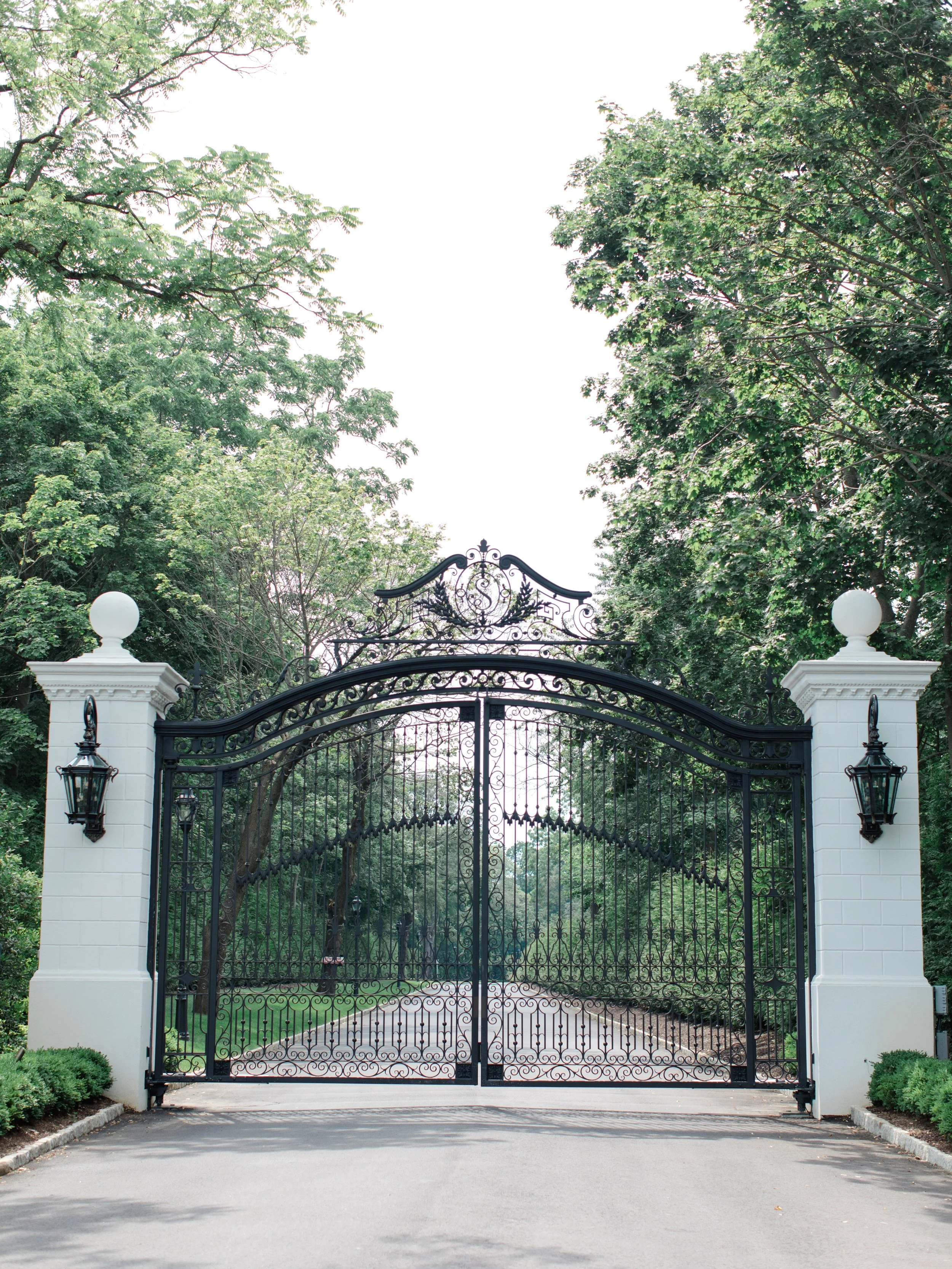 A decorative wrought iron gate flanked by white brick pillars with globe finials and lanterns, set in a lush green park or estate with trees and a paved pathway visible through the gate.