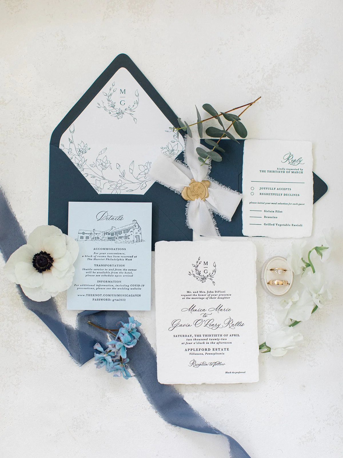 Wedding invitation suite with navy blue and white accents, rings, greenery, flowers, and RSVP card arranged on a white surface.