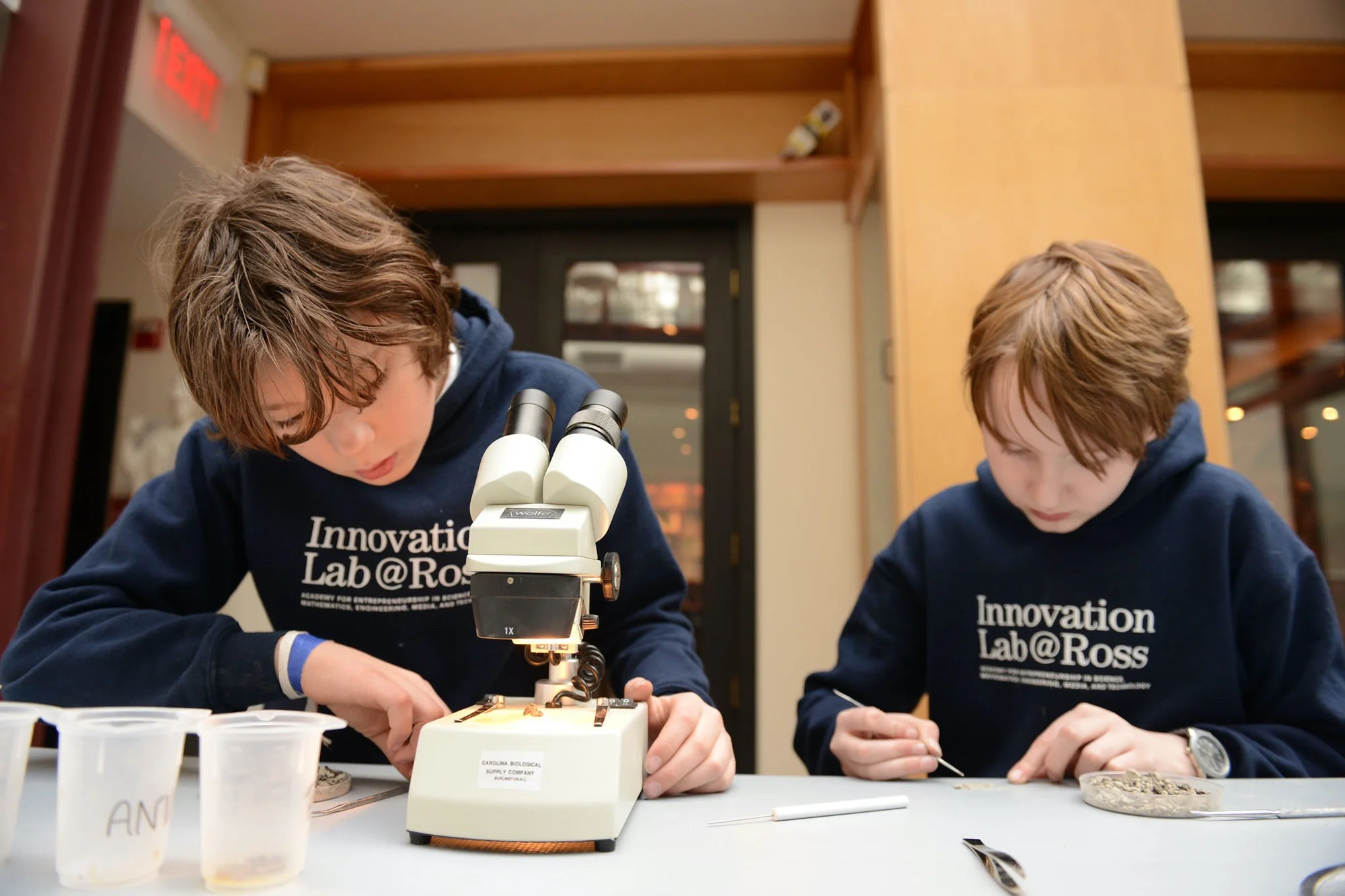 Two Ross STEAM Innovation Lab students seated at a table, one making adjustments to a microscope and the other preparing a specimen slide.