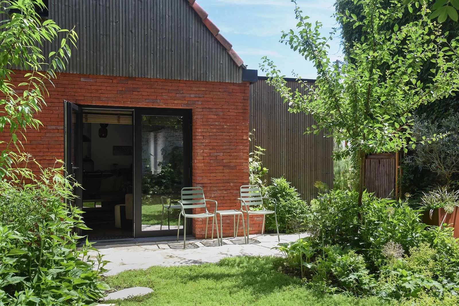 Landelijke tuin met weelderige beplanting tussen een bakstenen woning en een houten schuur