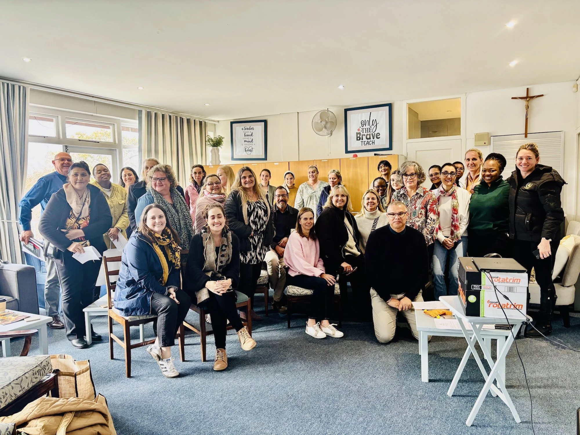 A group of diverse adults posing together in a room, smiling for a photo, with sunlight coming through windows. There are chairs, tables, and signs on the wall, including a crucifix and motivational quotes.
