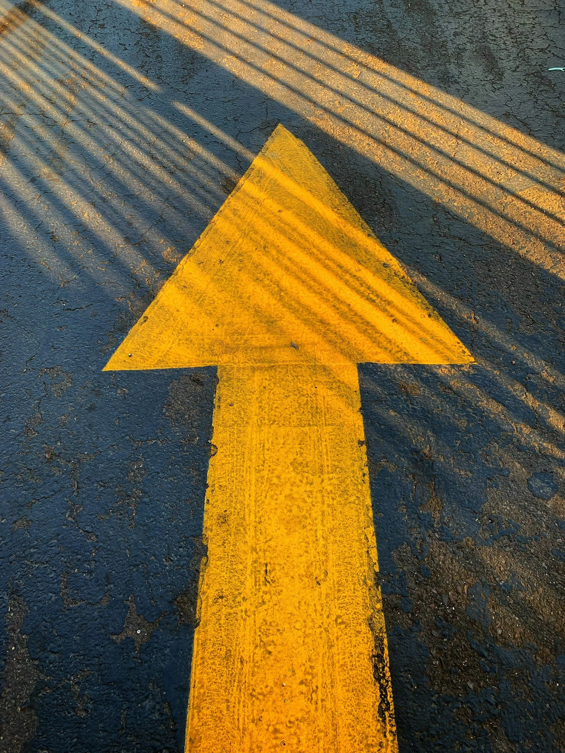 Yellow painted arrow on dark asphalt pointing forward, with shadows of railings or bars casting lines across the scene.