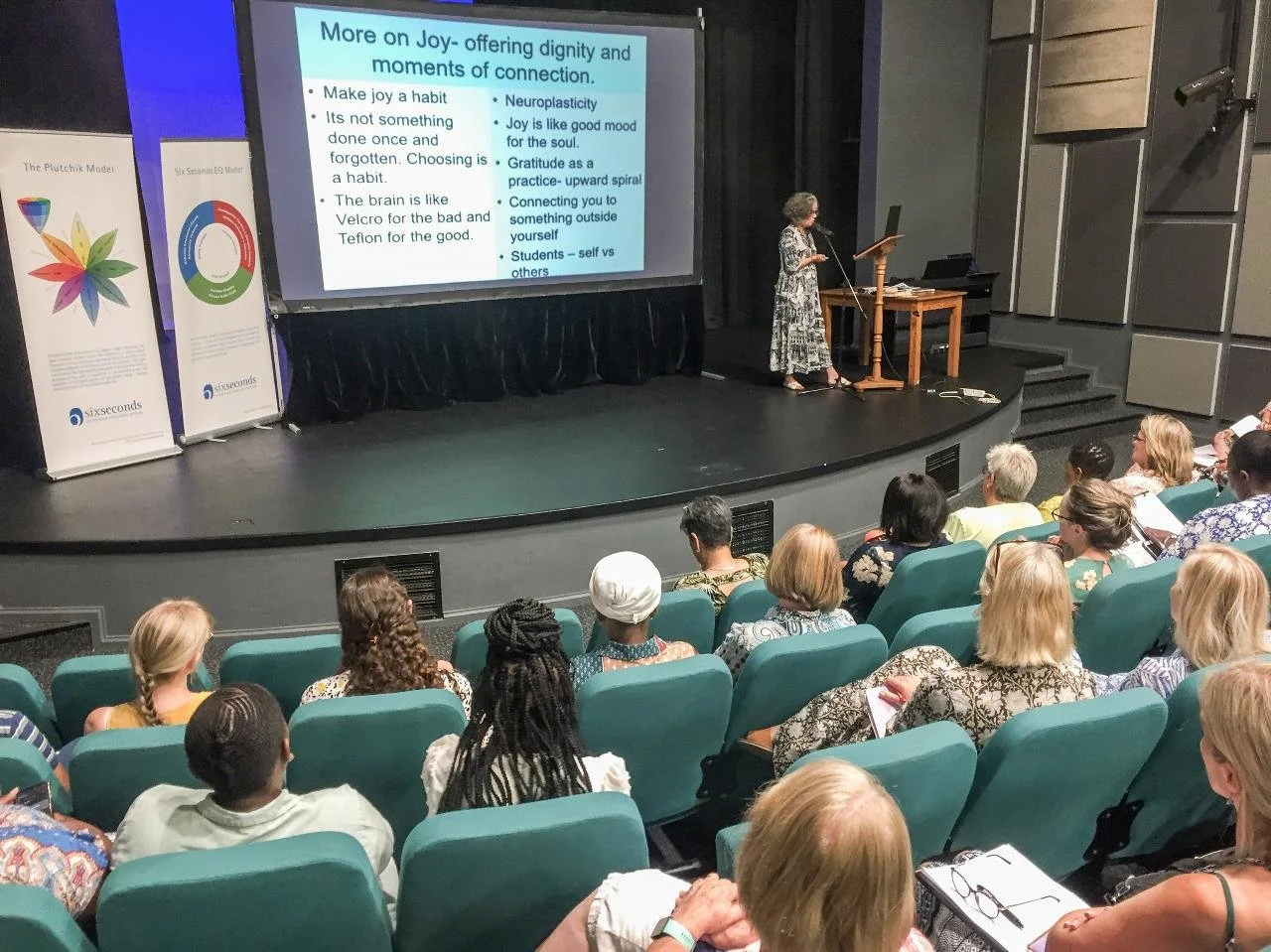 Libby Edmonds giving a presentation in a room with large windows. A large screen displays a slide about "Straw and Sticks in Modern Life" with text and illustrations of a brick wall, sticks, and hay.