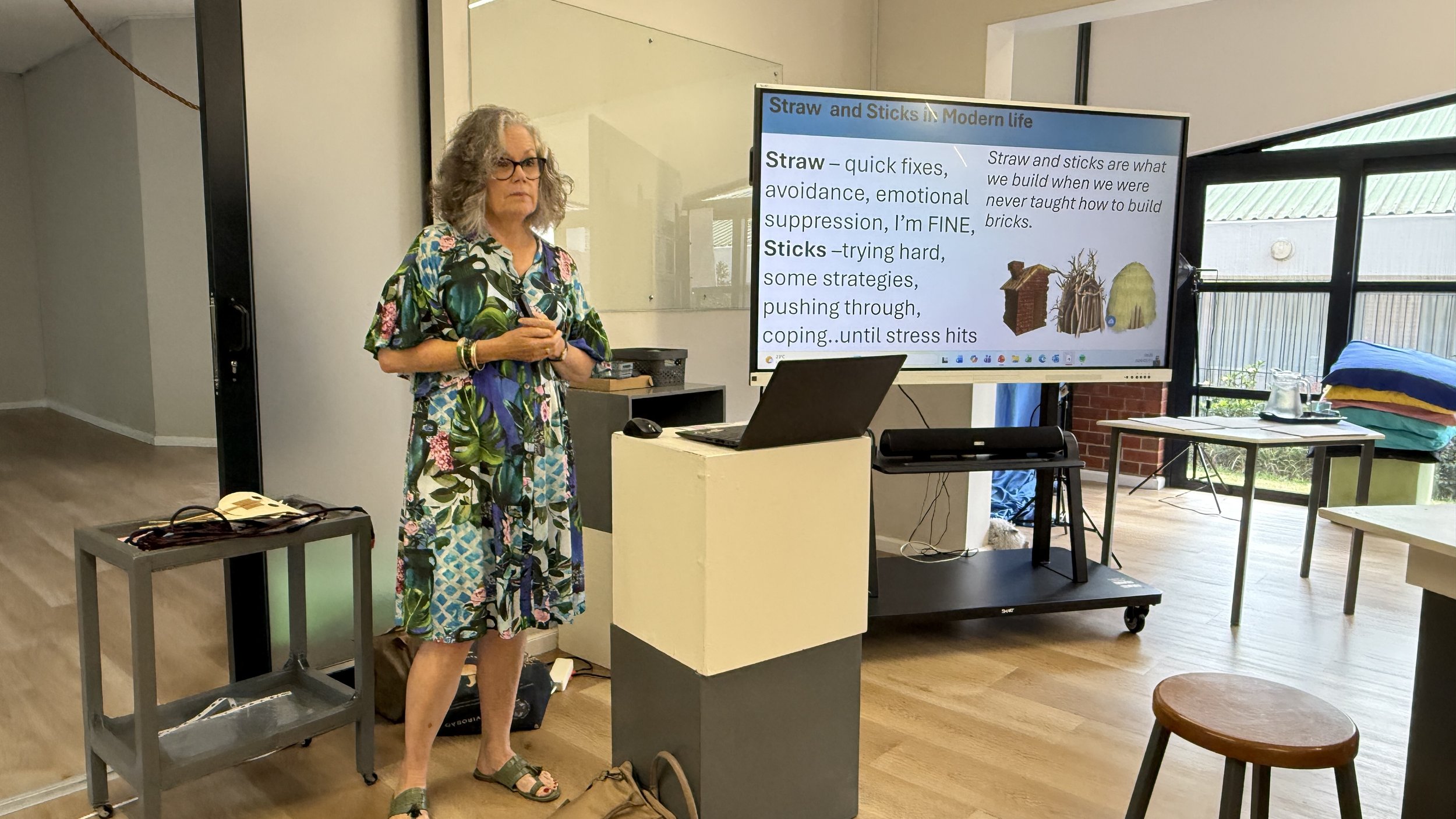 A woman with gray curly hair, glasses, wearing a colorful floral dress, stands in front of a presentation screen in a room with large windows. The screen displays a slide about