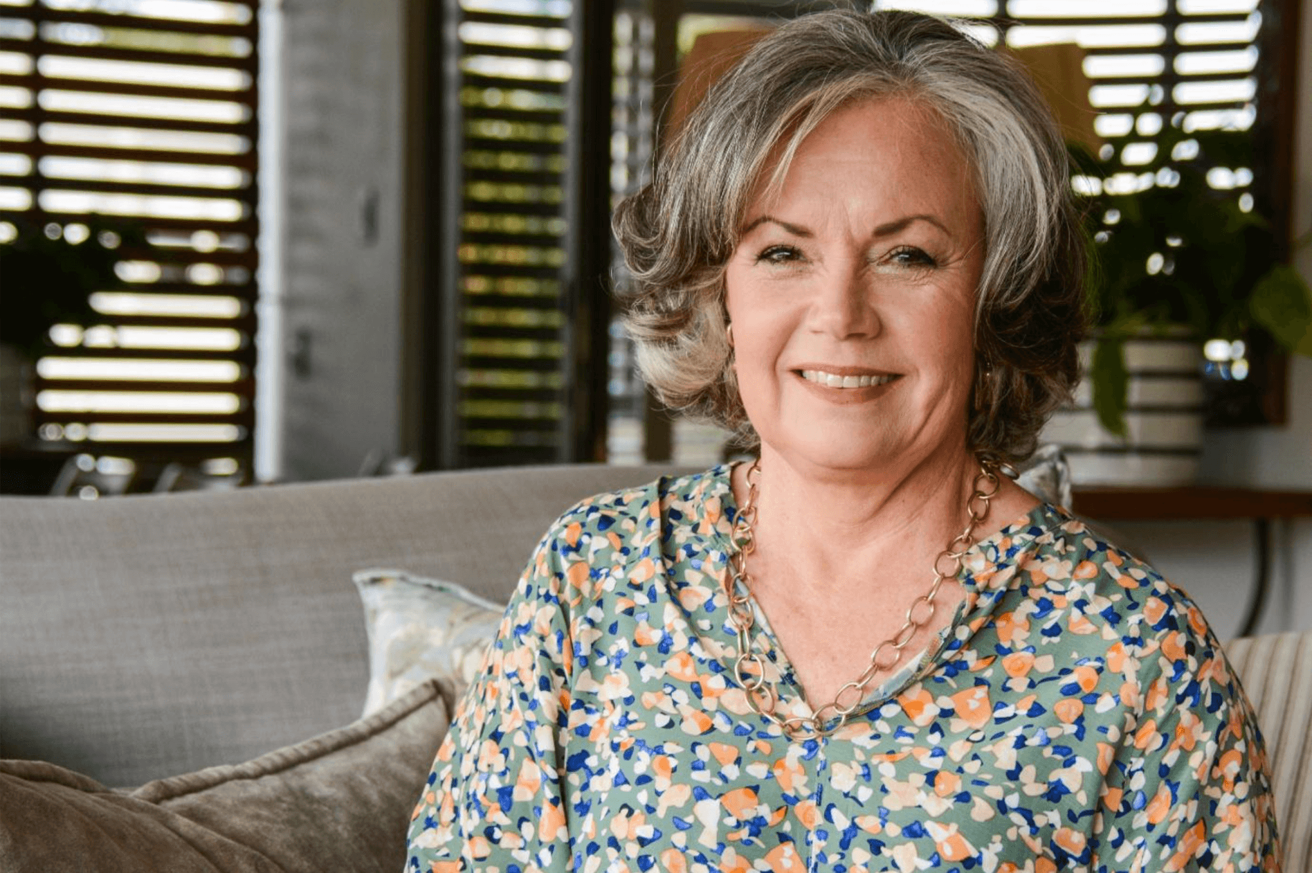 A smiling middle-aged woman with curly gray hair sitting on a sofa in a cozy living room with wooden window blinds and houseplants in the background.