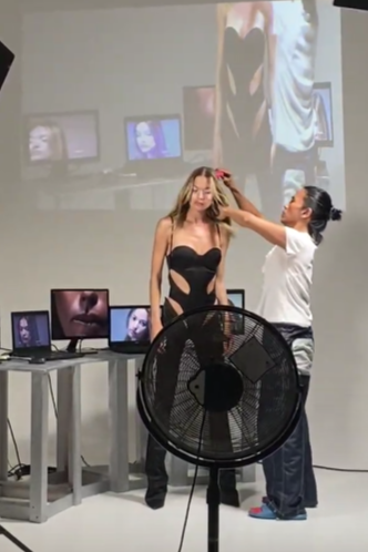 A woman in a black dress is having her hair styled by a hairstylist in a room with multiple computer monitors, a large fan, and a projected image on the wall.