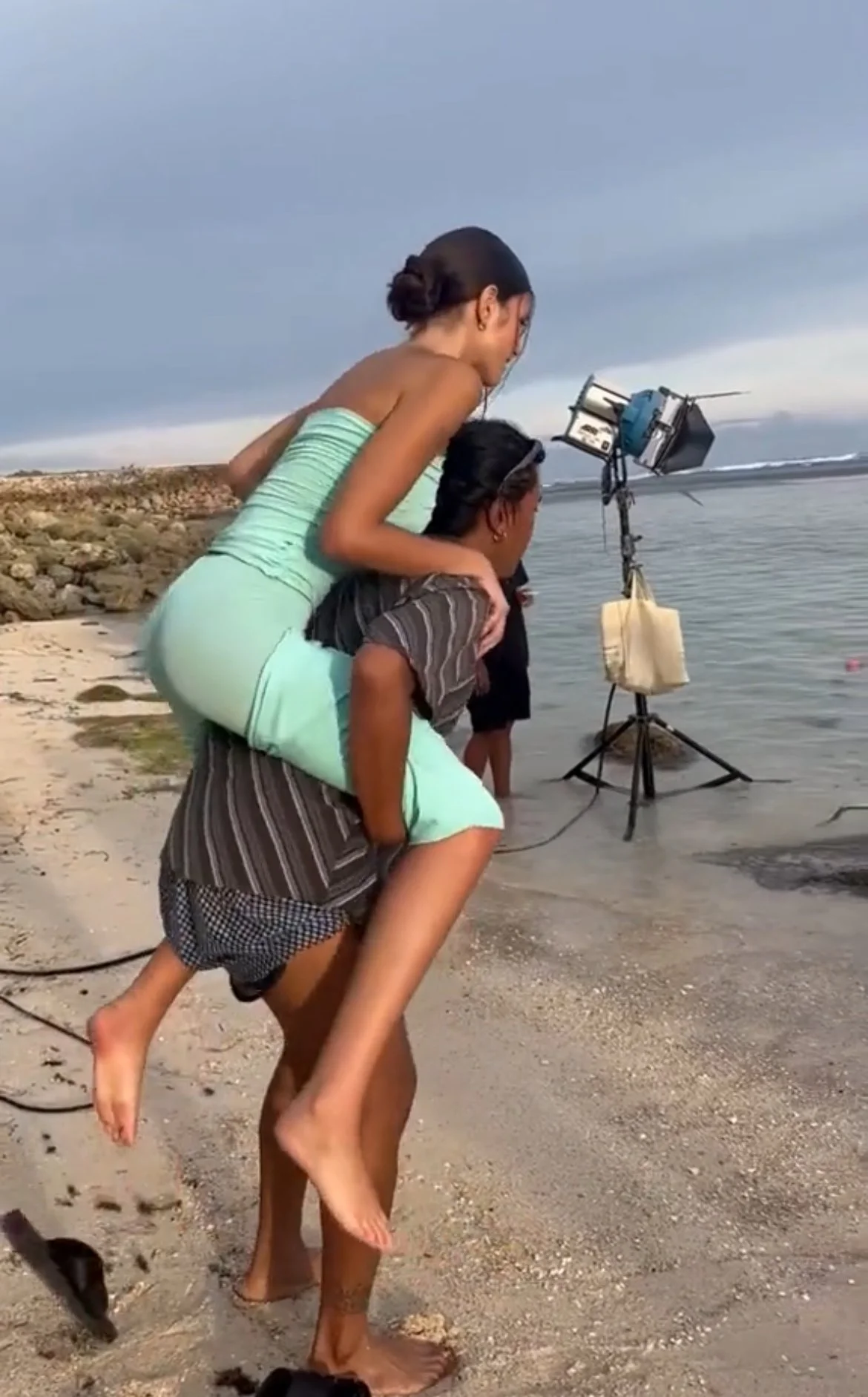Two young women are on a beach, one giving the other a piggyback ride. They are near a camera setup on a tripod, with equipment attached, and a cloudy sky overhead.
