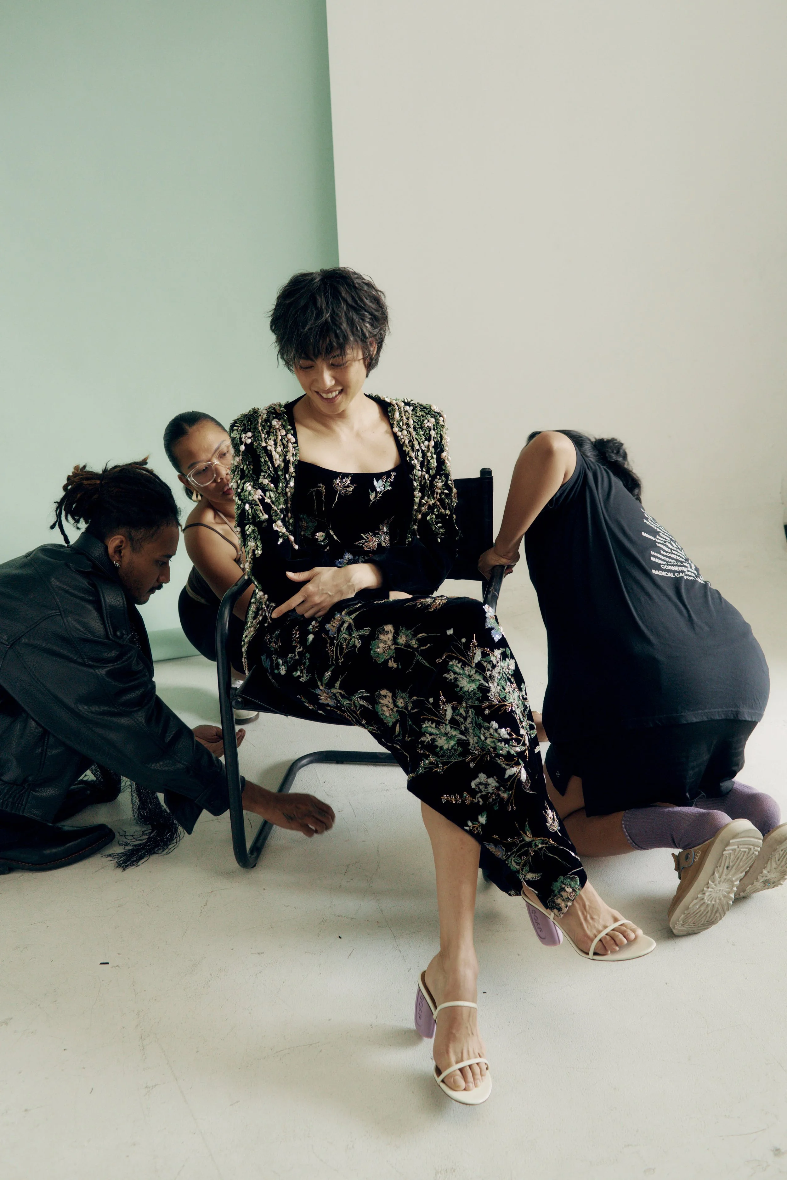 A woman in a floral dress and white high-heeled sandals is seated on a chair, smiling with her eyes closed. She is surrounded by four people assisting her, with one person adjusting her dress, two kneeling nearby, and another person’s head and hand visible. The setting is a plain white room.