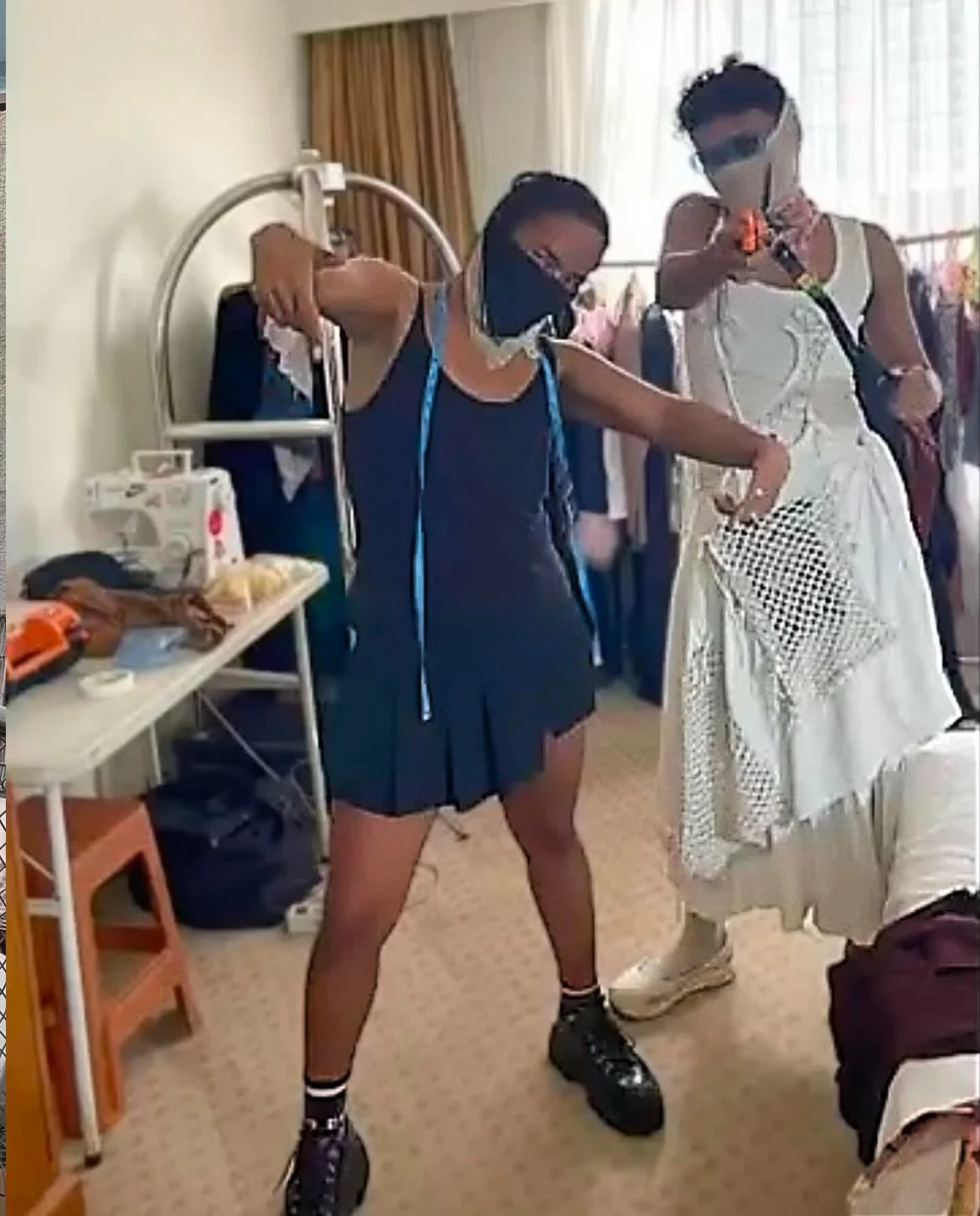 A young woman wearing a black face mask, black shorts, black sneakers, and a black tank top stands in a hospital room. She is taking a selfie with her phone, while another woman in a white dress and black sunglasses poses nearby. The room has hospital equipment and supplies on a table.