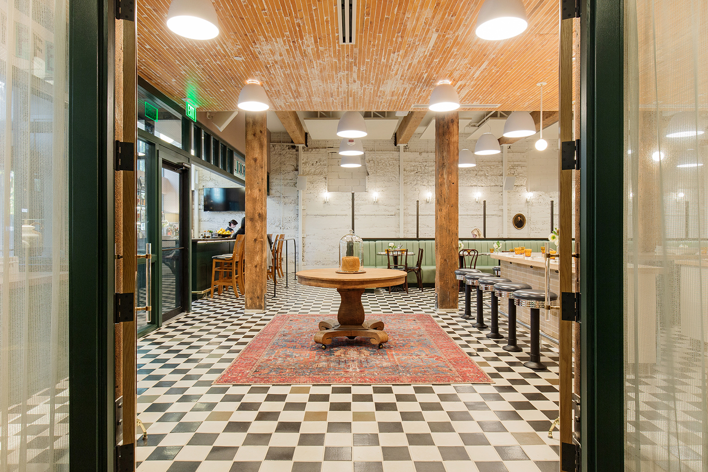Shot from entry looking into restaurant. Checker patterned floor, large round wooden table on ornamental rug, bar and booth seating also pictured.