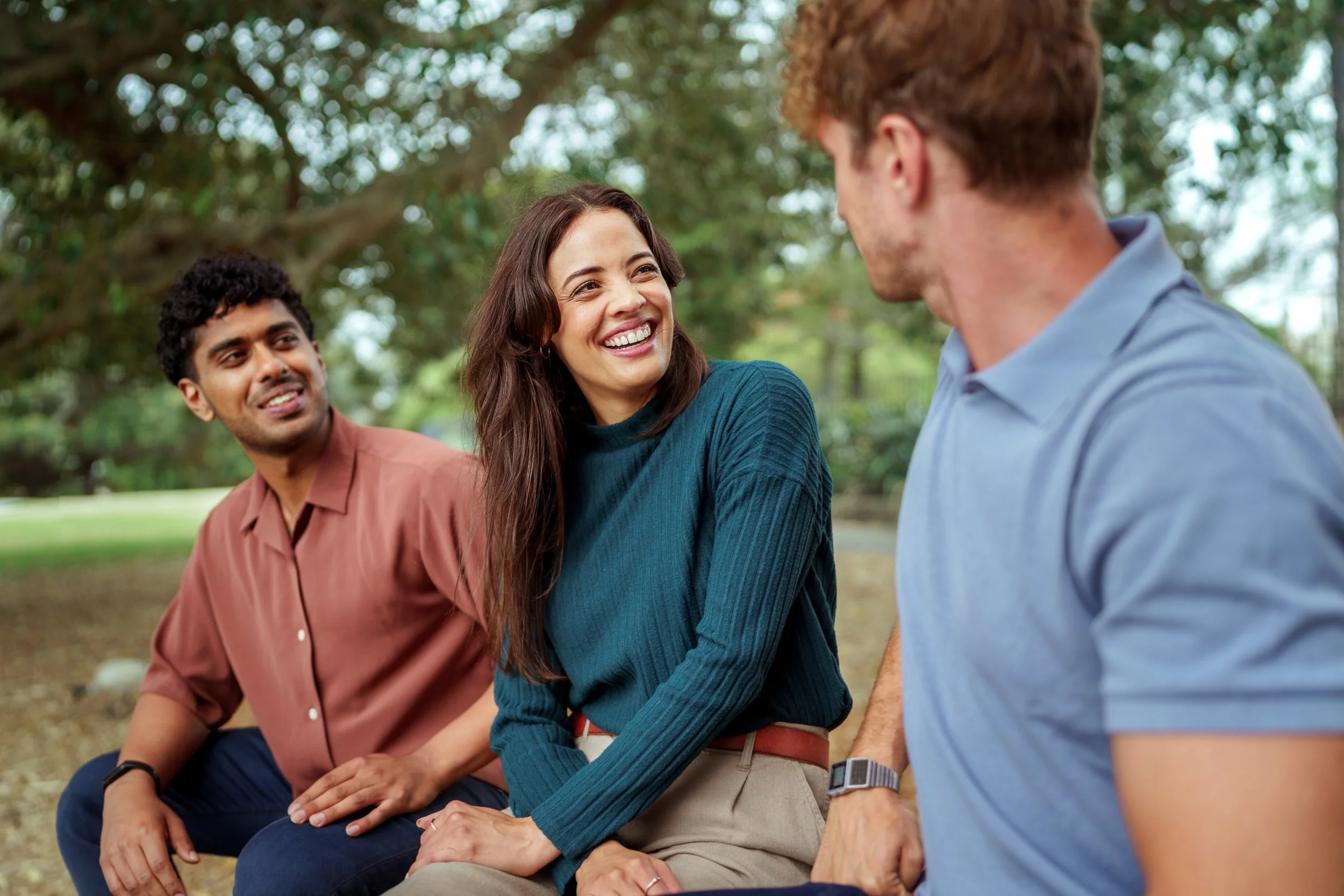 Two smiling young people sitting in a park looking at another person out of frame