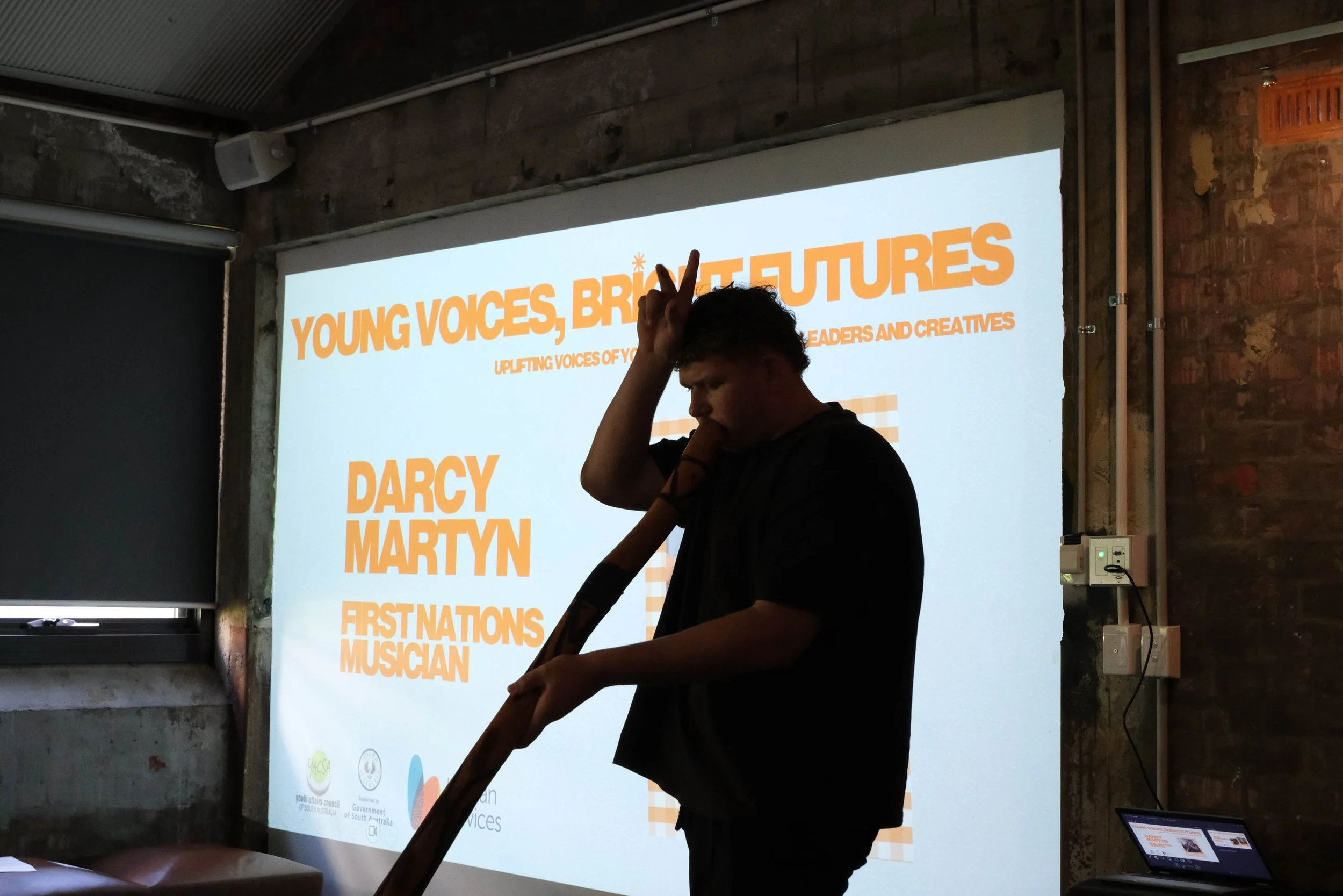 Darcy plays yidaki with his right hand raised to form ears on his head with his fingers