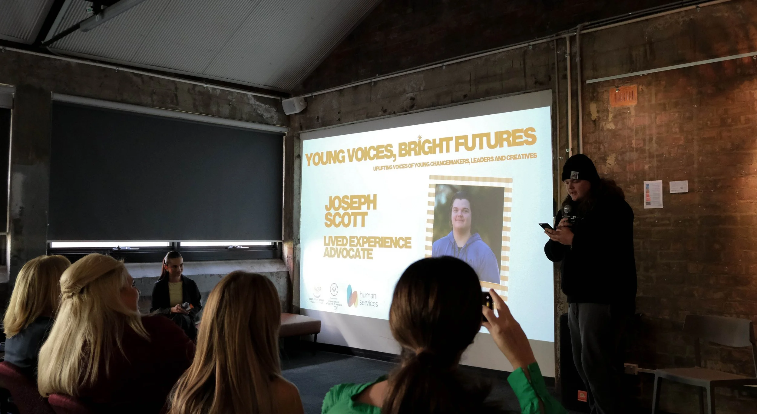 Joseph speaks to audience in front of projector screen