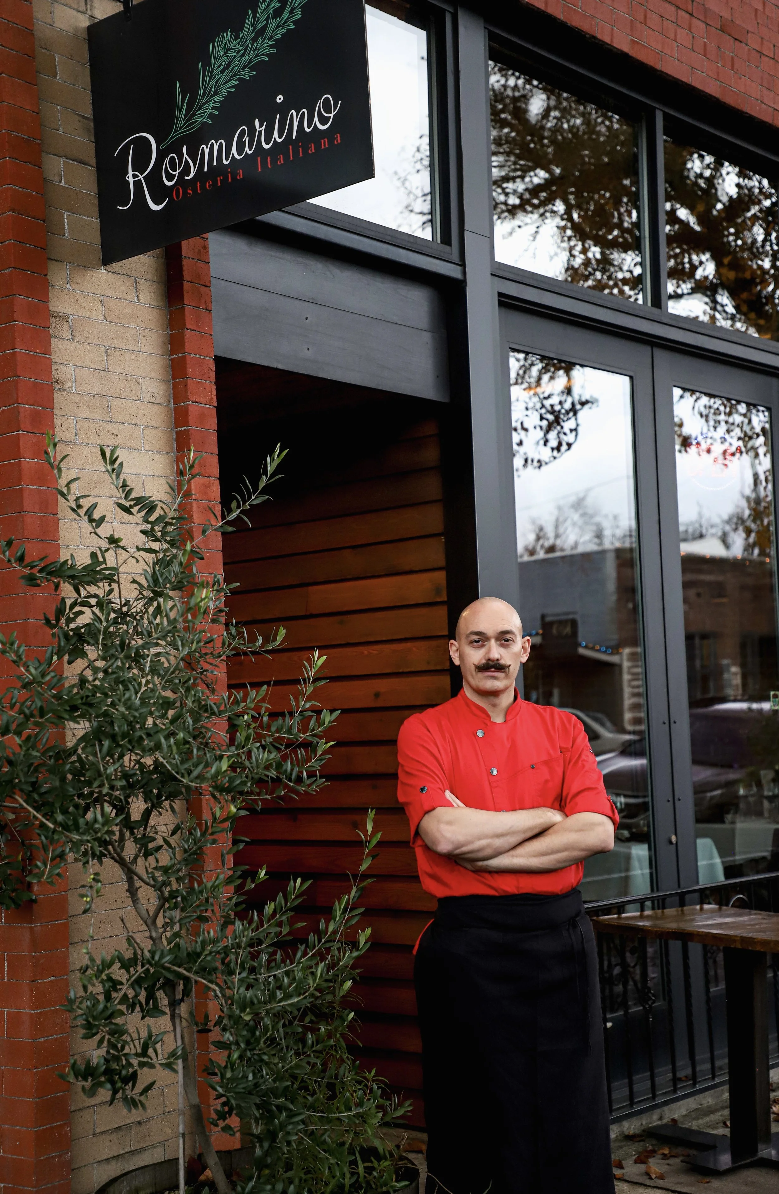 A chef with a mustache, wearing a red shirt and black apron, standing outside a restaurant called Rosmarino Osteria Italiana, with arms crossed in front of a wooden wall and glass windows.