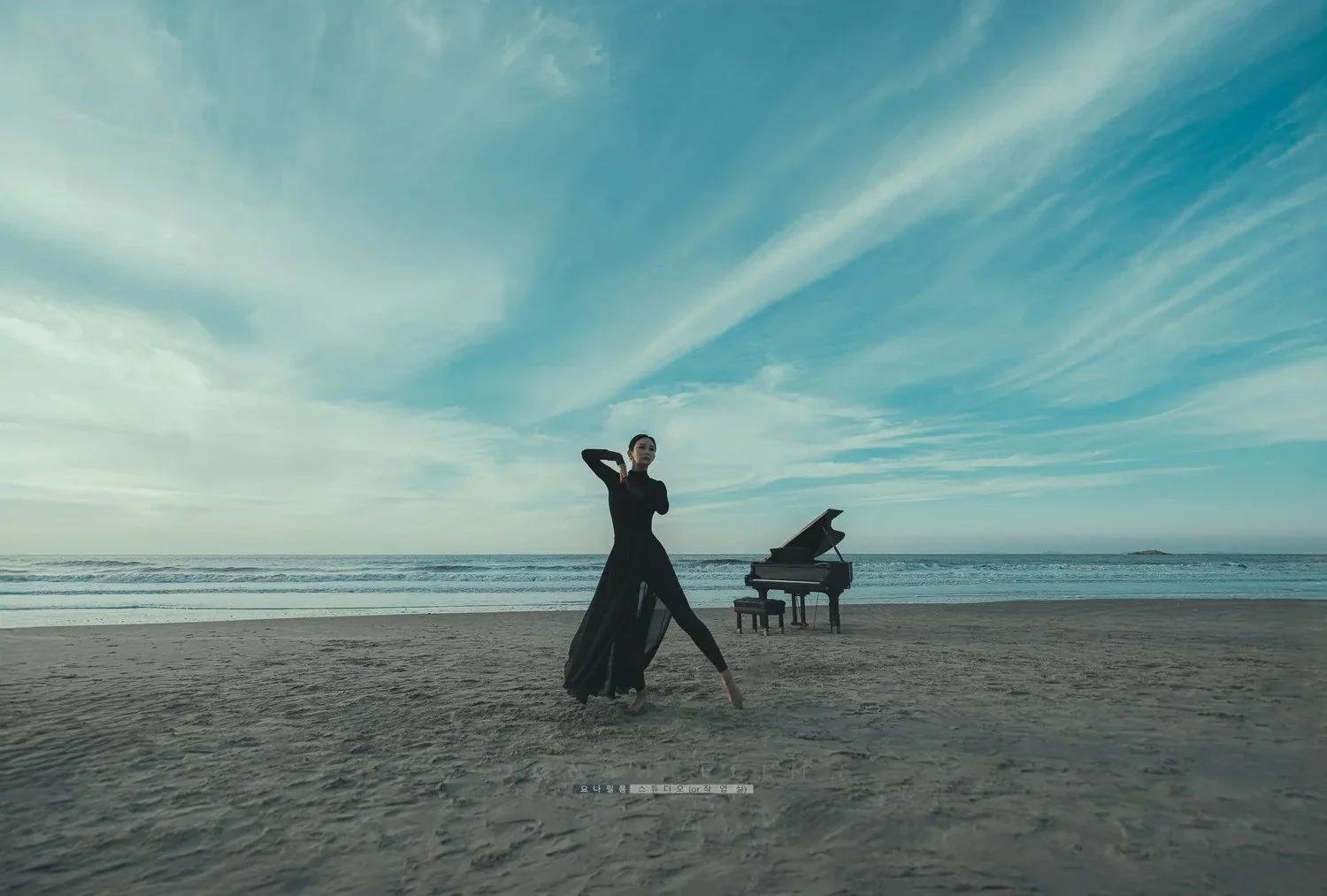 A woman dressed in a black flowing dress standing on a sandy beach near the ocean with a grand piano nearby and a blue sky with wispy clouds above.