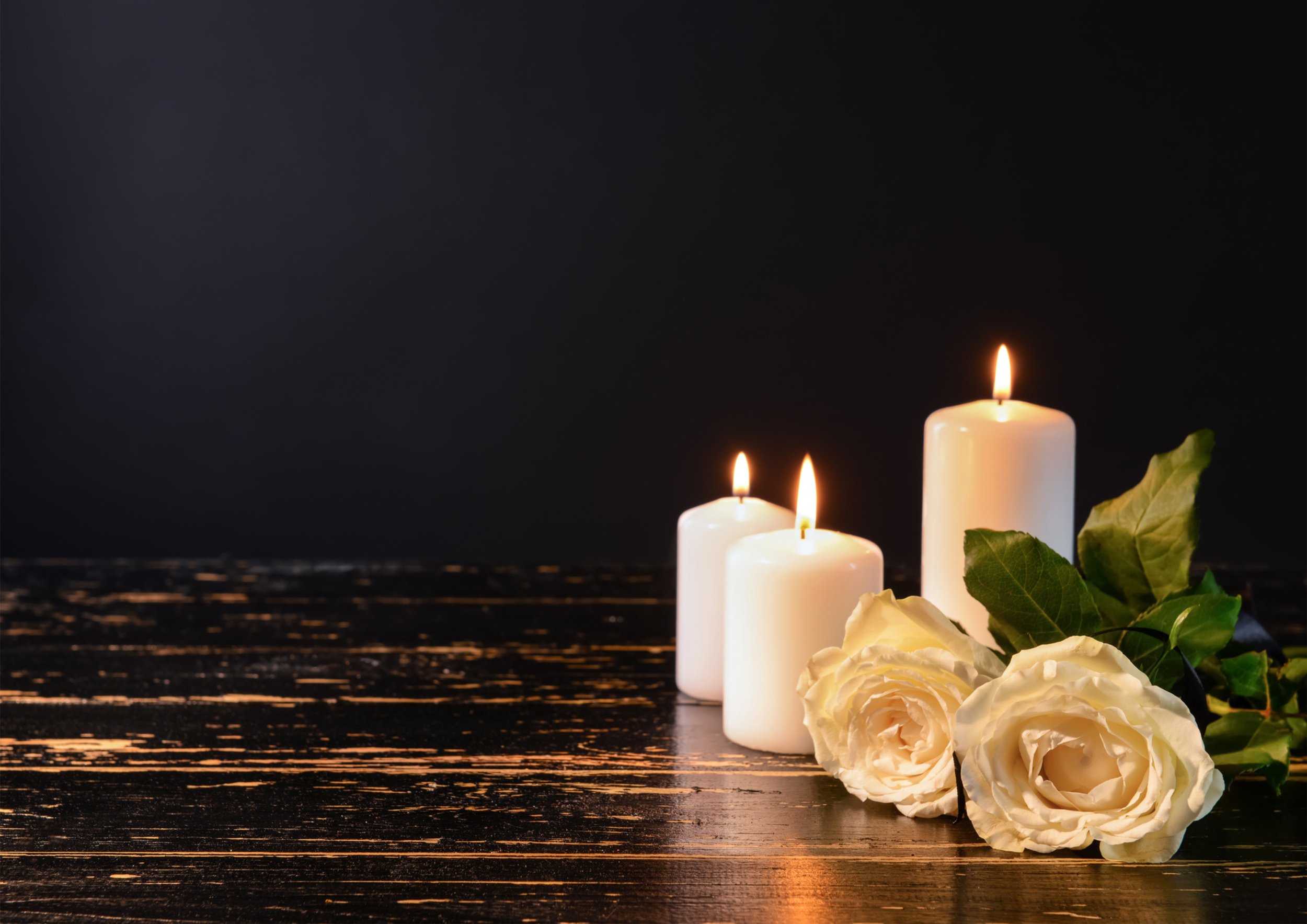 Three lit white candles with green leaves and white roses on a dark wooden surface against a black background.
