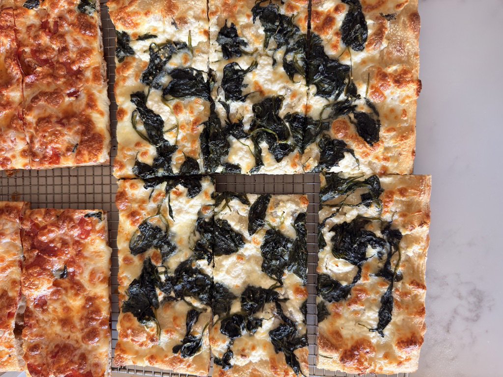 Four slices of flatbread pizza with black leafy toppings, on a cooling rack.
